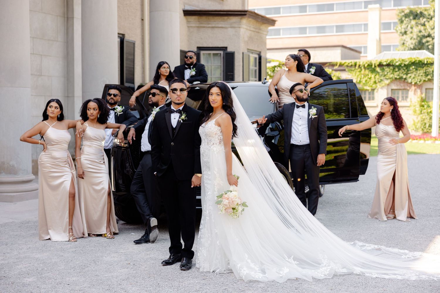 Wedding party in elegant champagne dresses and black tuxedos gathered outside historic venue.
