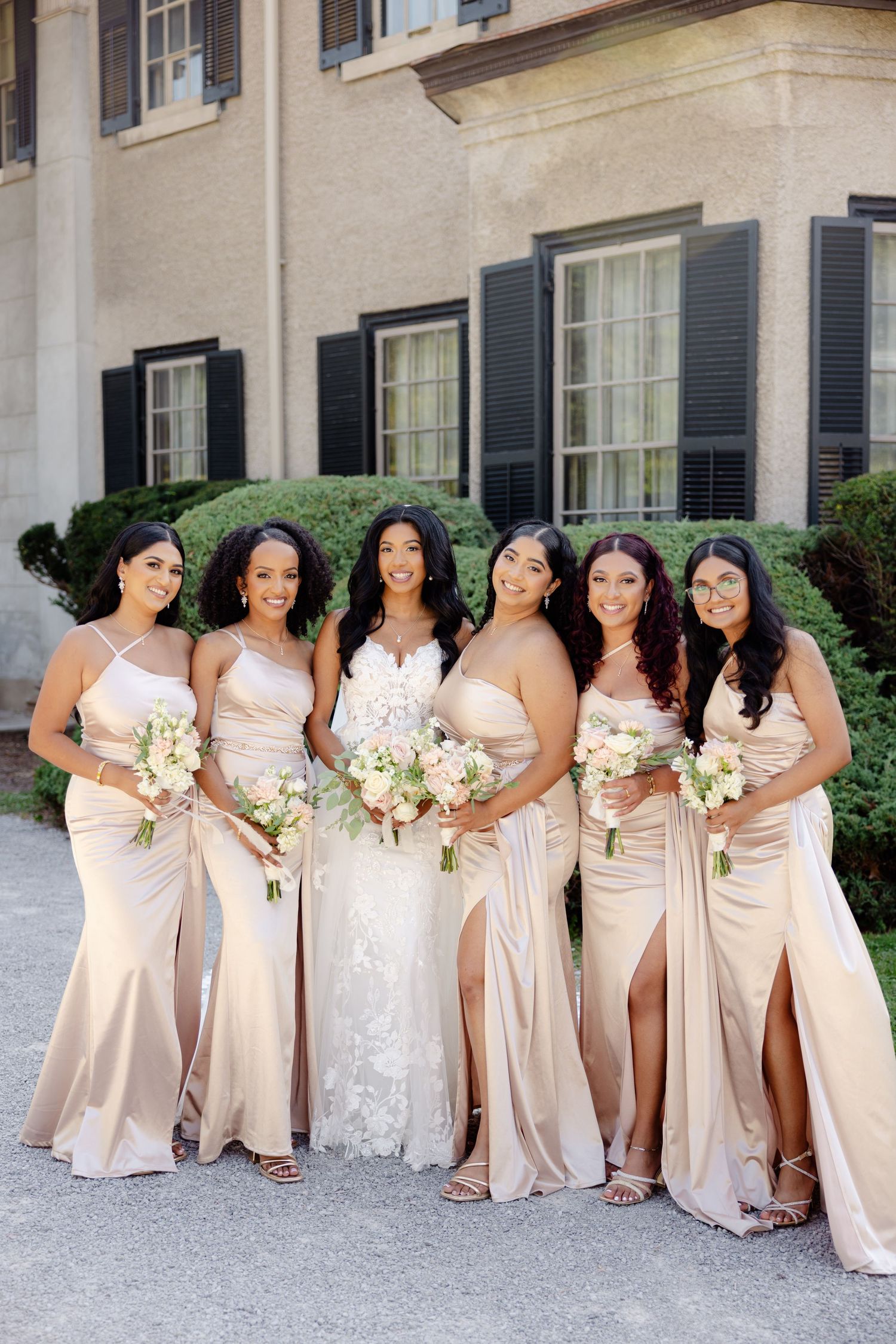 Bridal party in flowing champagne dresses poses with bouquets against classic architecture.