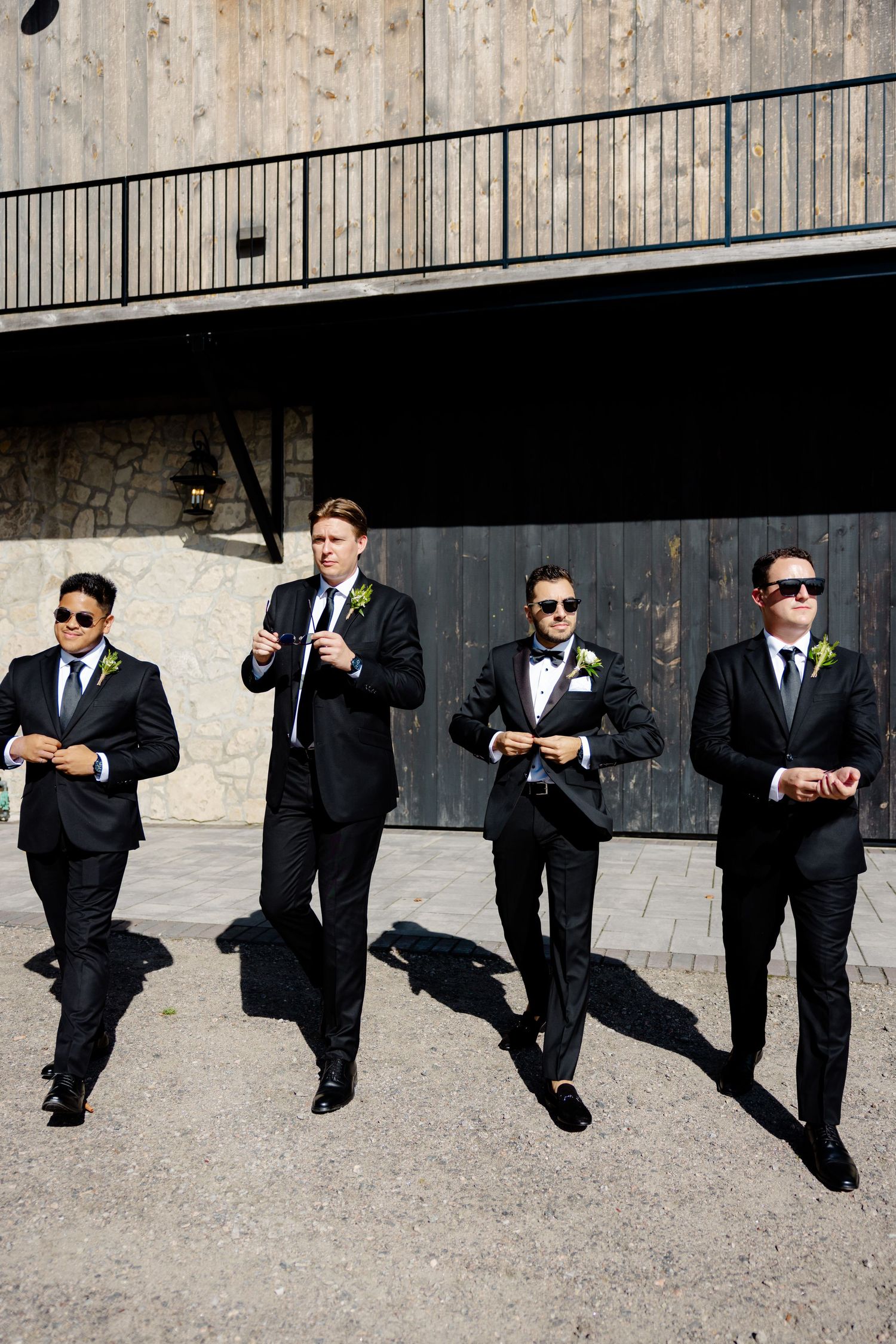 A group of groomsmen in black suits and sunglasses walk confidently in formation against a rustic backdrop.