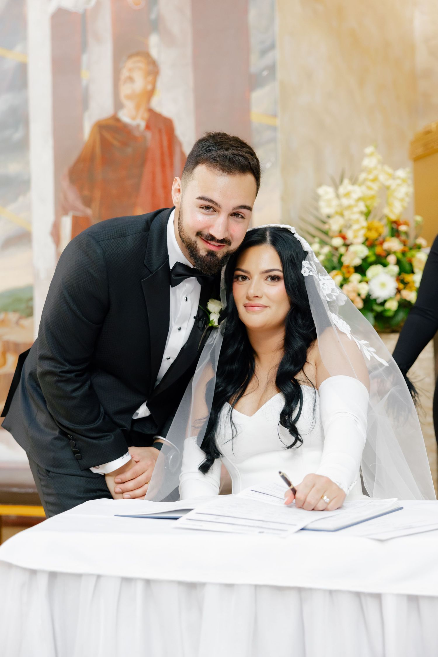 A couple signing their marriage license at a white-clothed table with floral decorations.