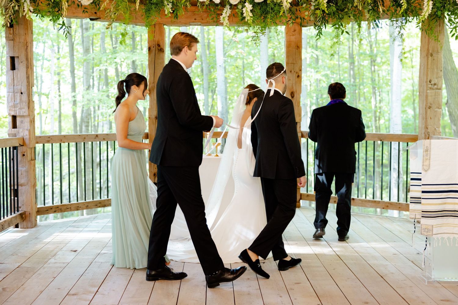 Wedding party gathering in rustic wooden pavilion with forest backdrop.