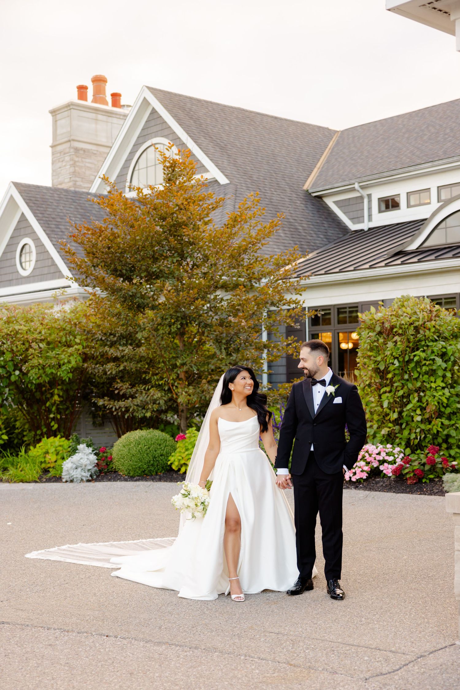 Newlyweds pose outside elegant grey house with fall foliage and architectural details.