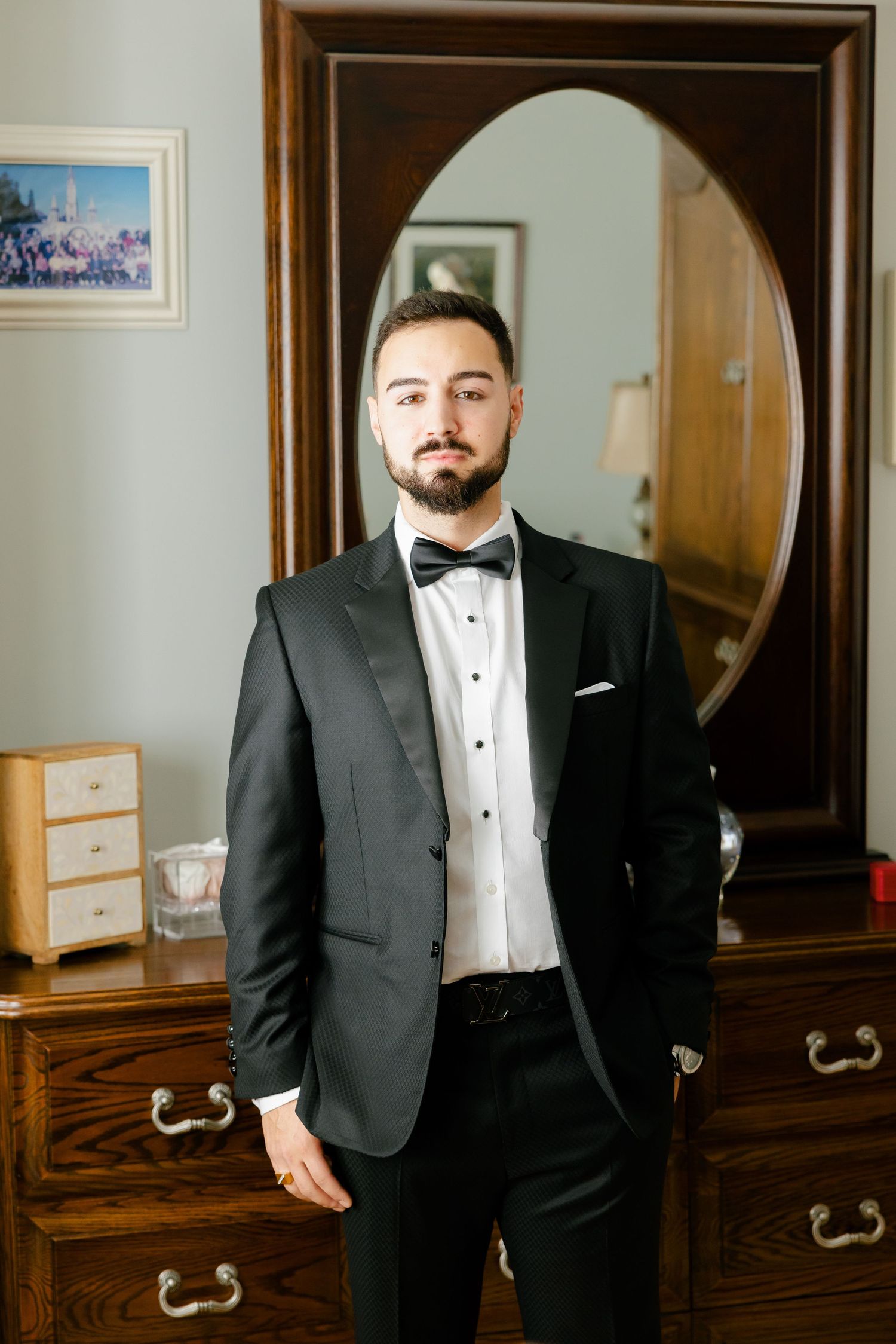 Classic black tuxedo with bow tie photographed against wooden furniture and oval mirror.