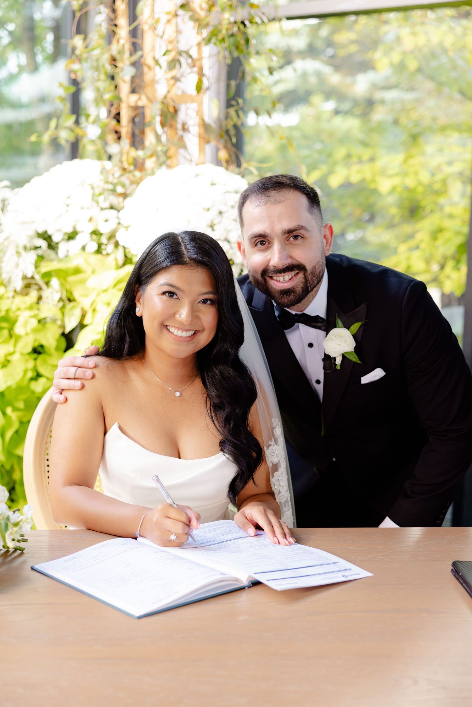 A couple signs their marriage license at a wooden table with greenery in the background.