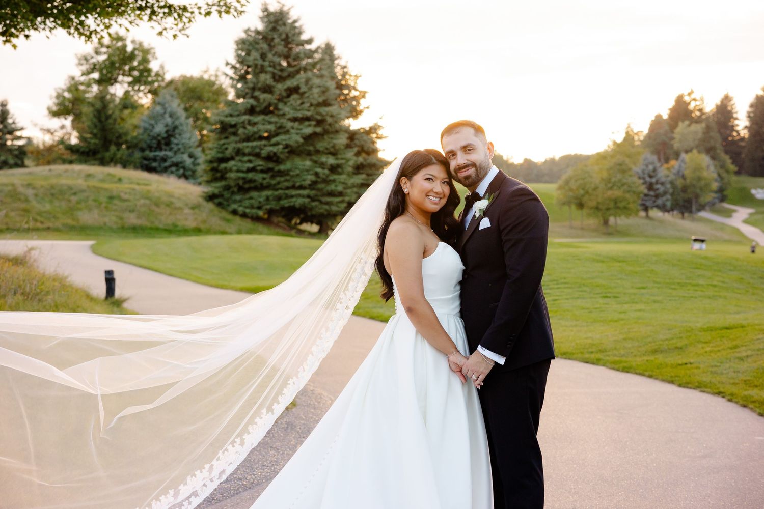 Newlyweds embrace on golf course path with flowing veil at sunset.