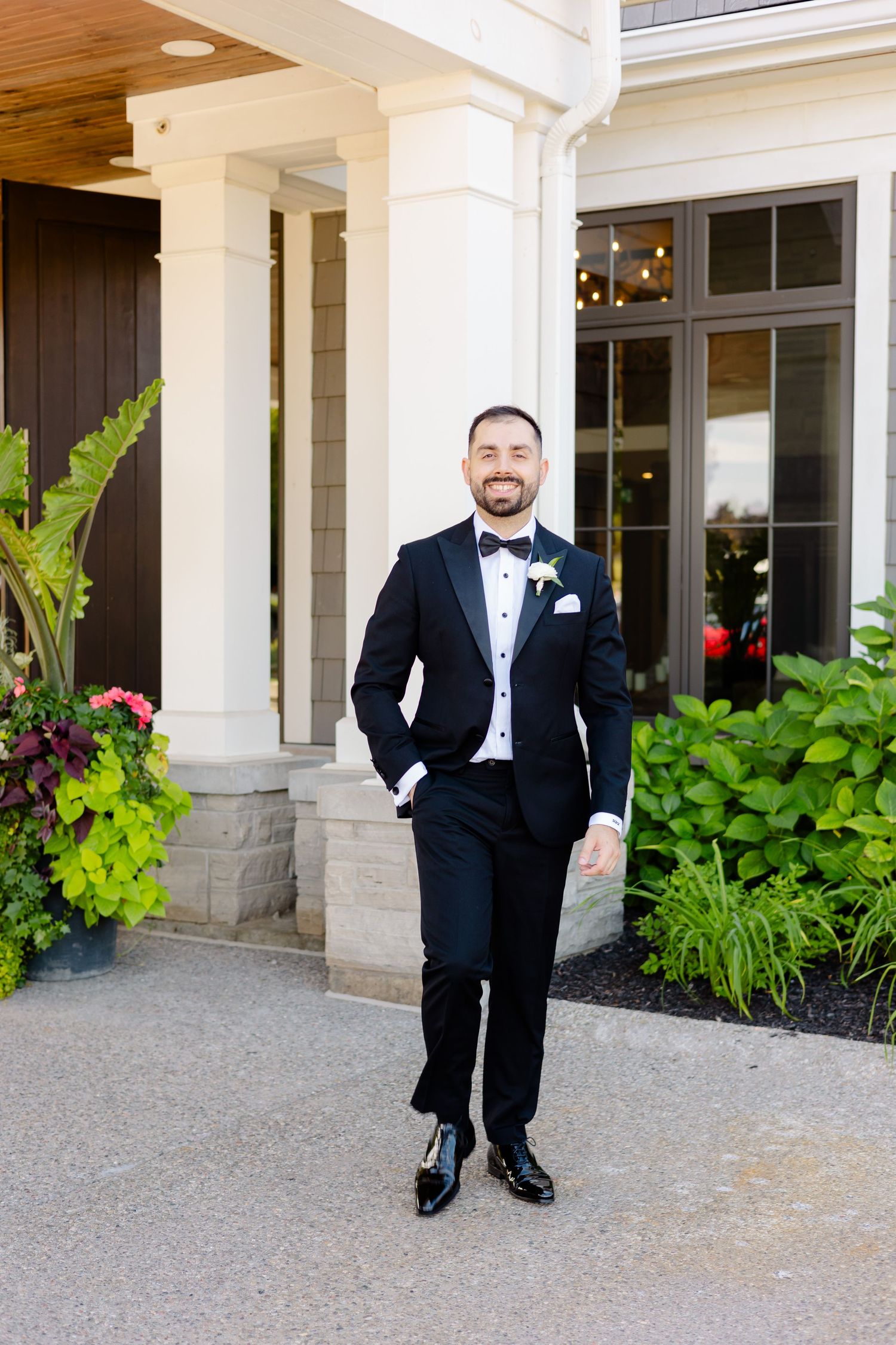 A formal portrait of someone in a classic black tuxedo standing on a porch with white columns and plants.