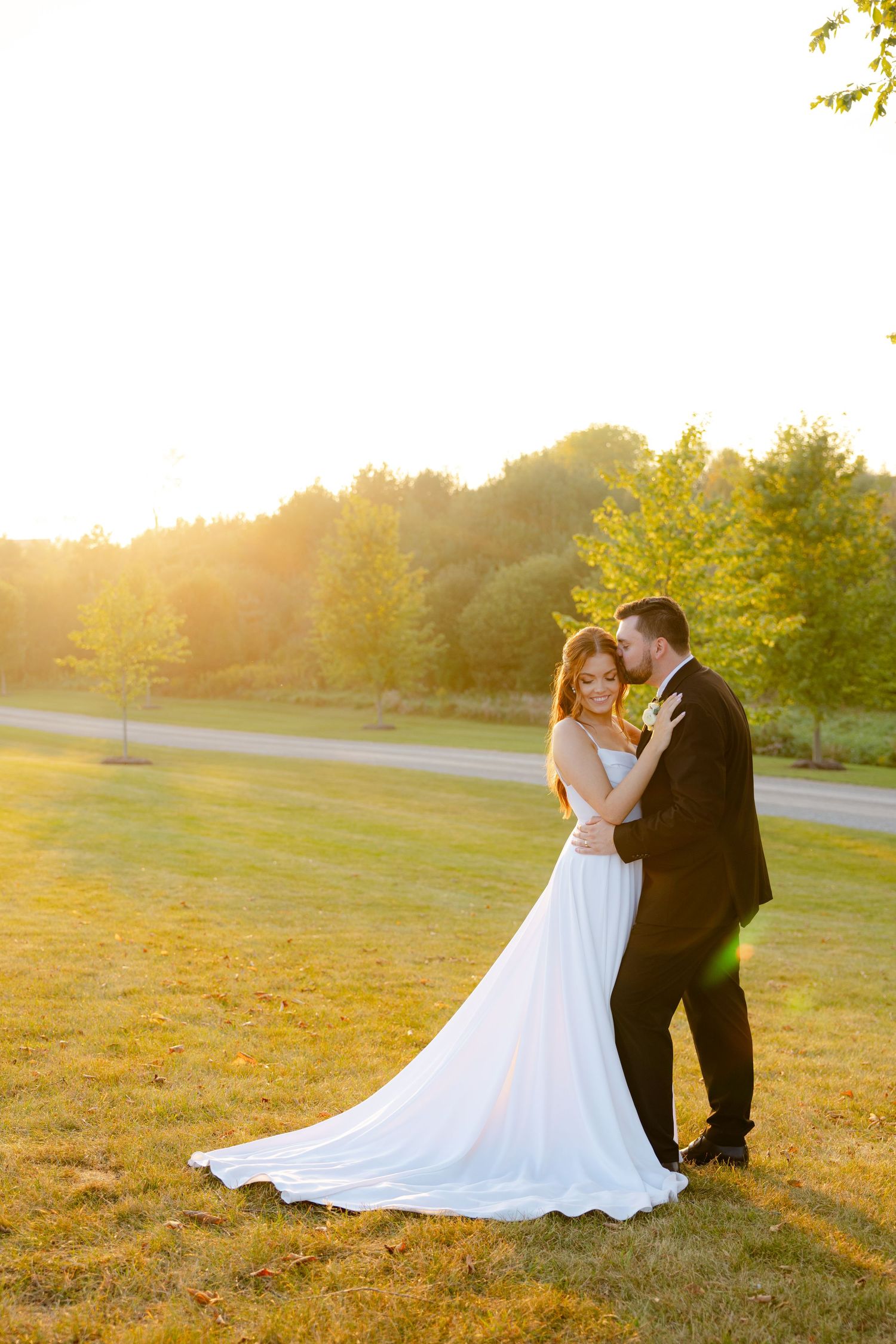 A romantic wedding couple embraces during golden hour sunset in a grassy field with trees in the background.