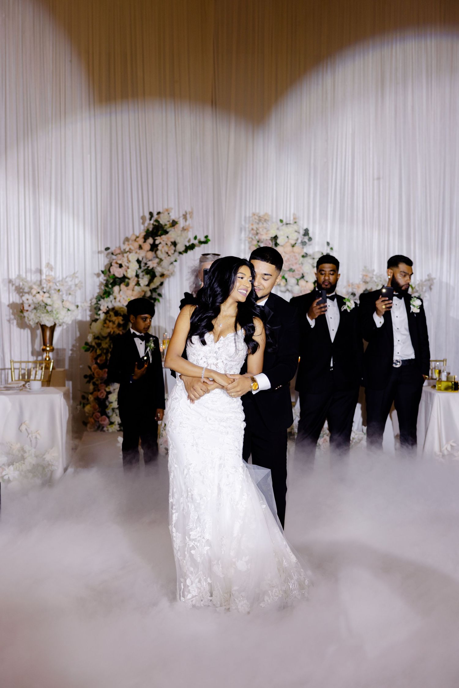 A couple shares an intimate dance moment at their wedding reception with guests and misty effects.