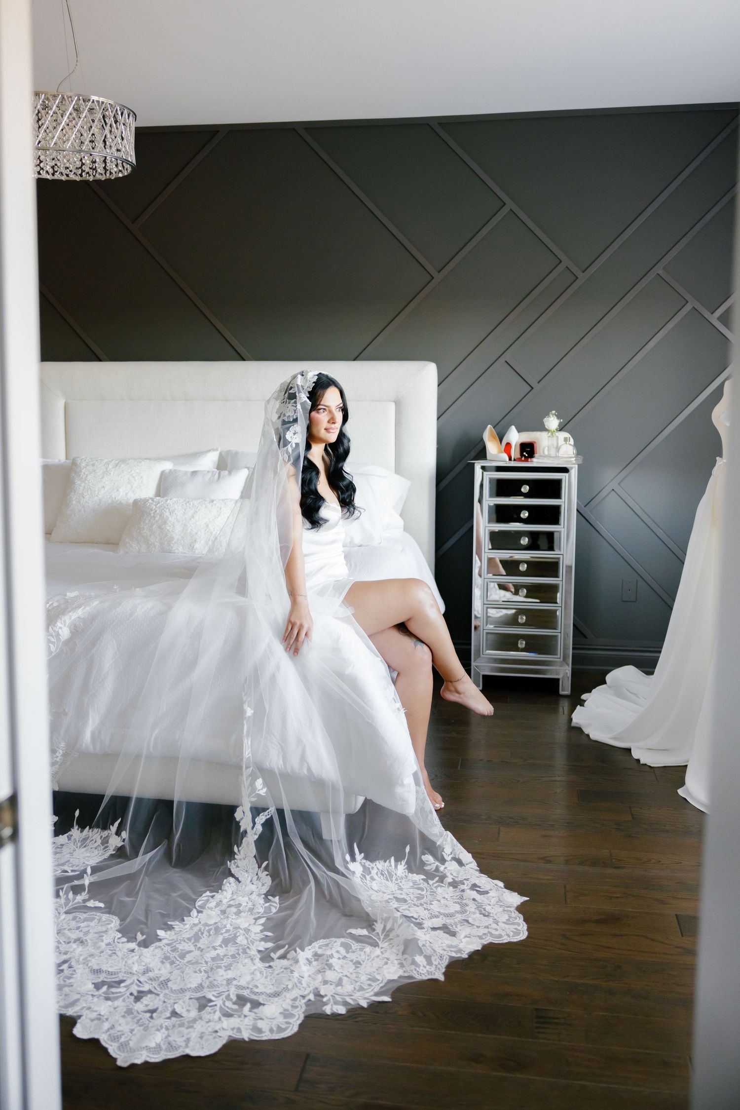 A bride in a white lace gown with long veil sits on a white bed against a dark geometric wall.