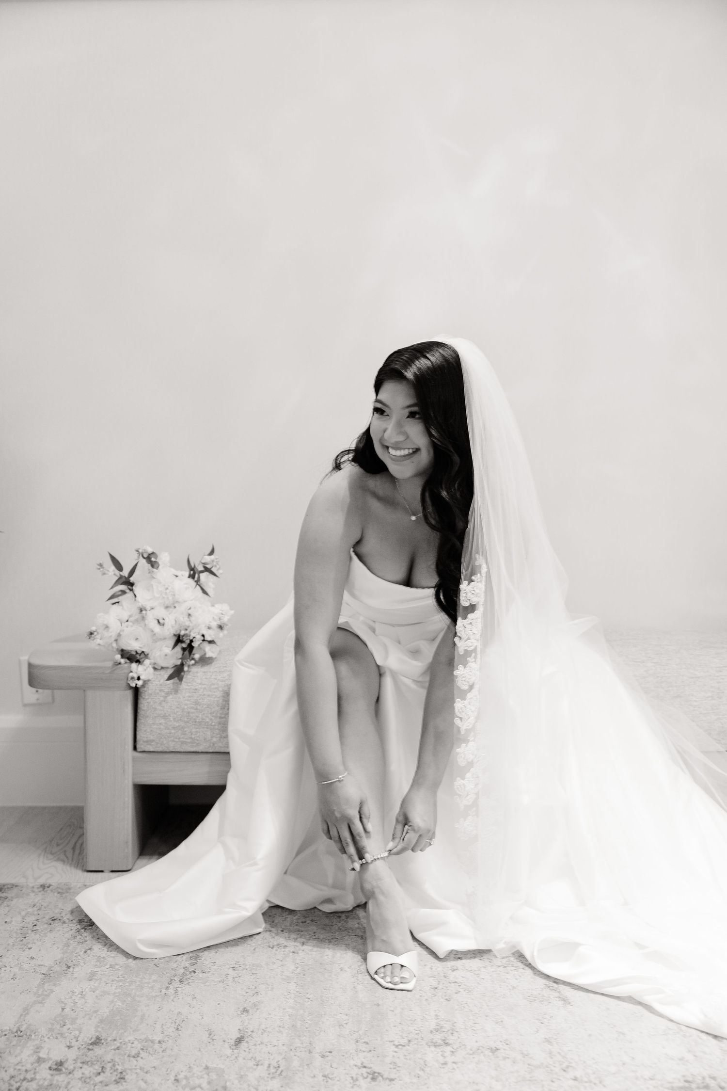 Black and white portrait of bride adjusting her shoe with floral arrangement in background.