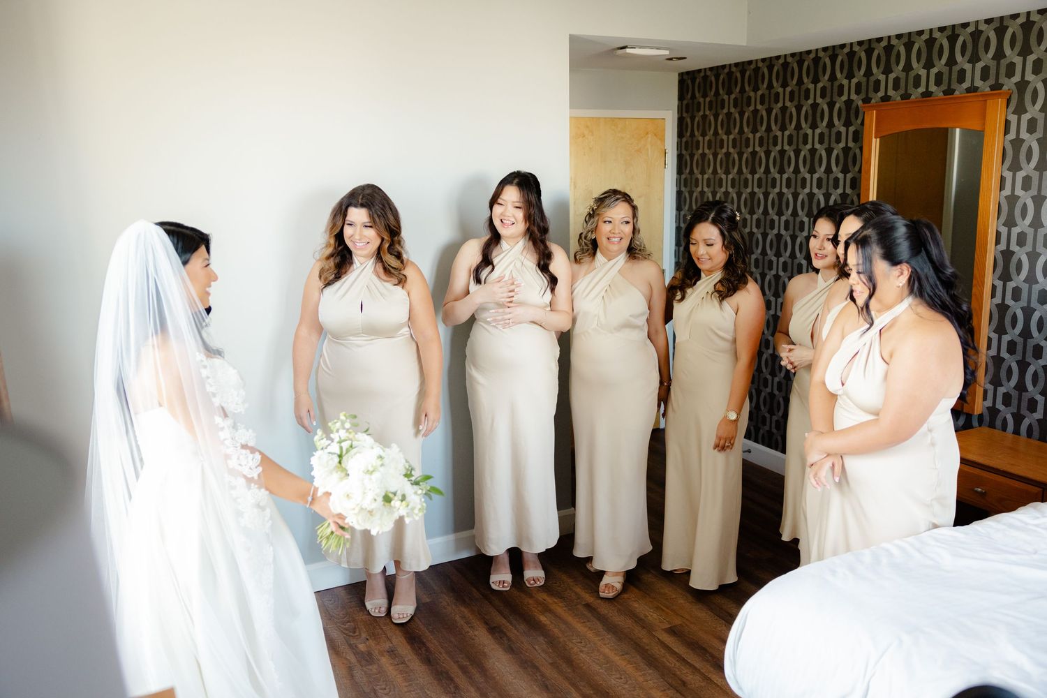 Bridesmaids in elegant cream-colored dresses gather in a hotel room before the wedding ceremony.