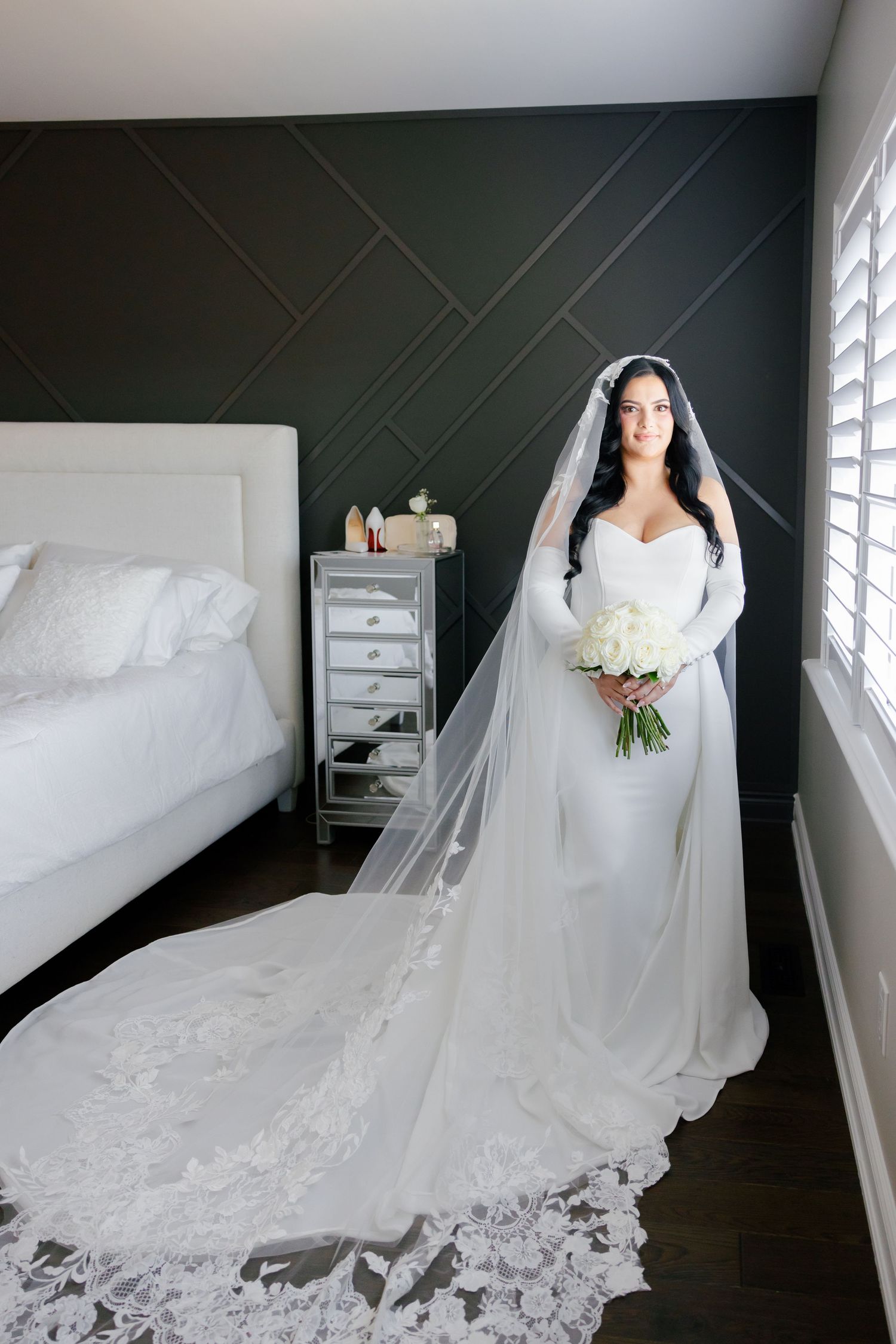 Bride in fitted wedding gown with dramatic cathedral train poses by bedroom window.
