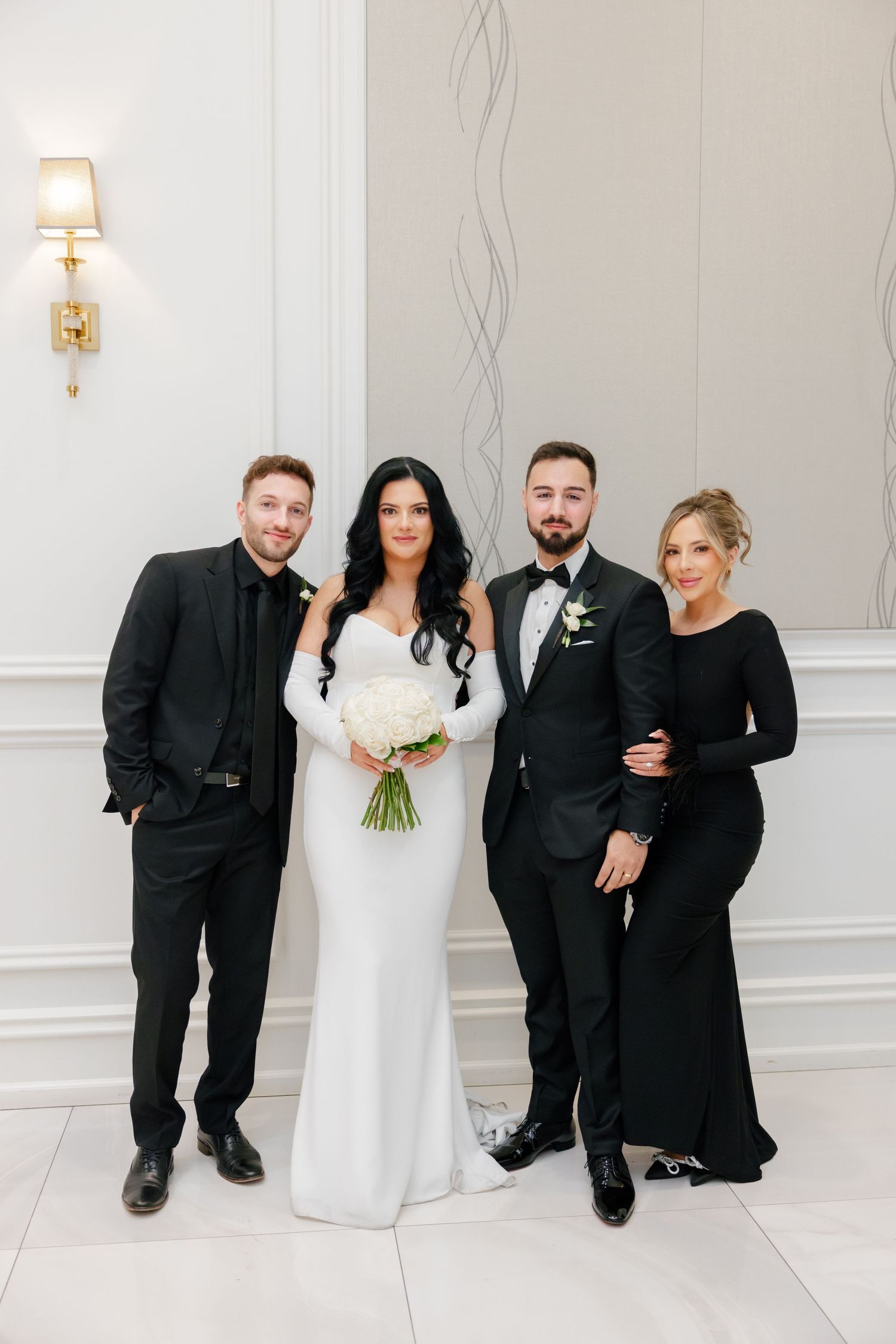 Wedding party poses against white wall wearing elegant black attire with bride in sleek white dress holding bouquet.