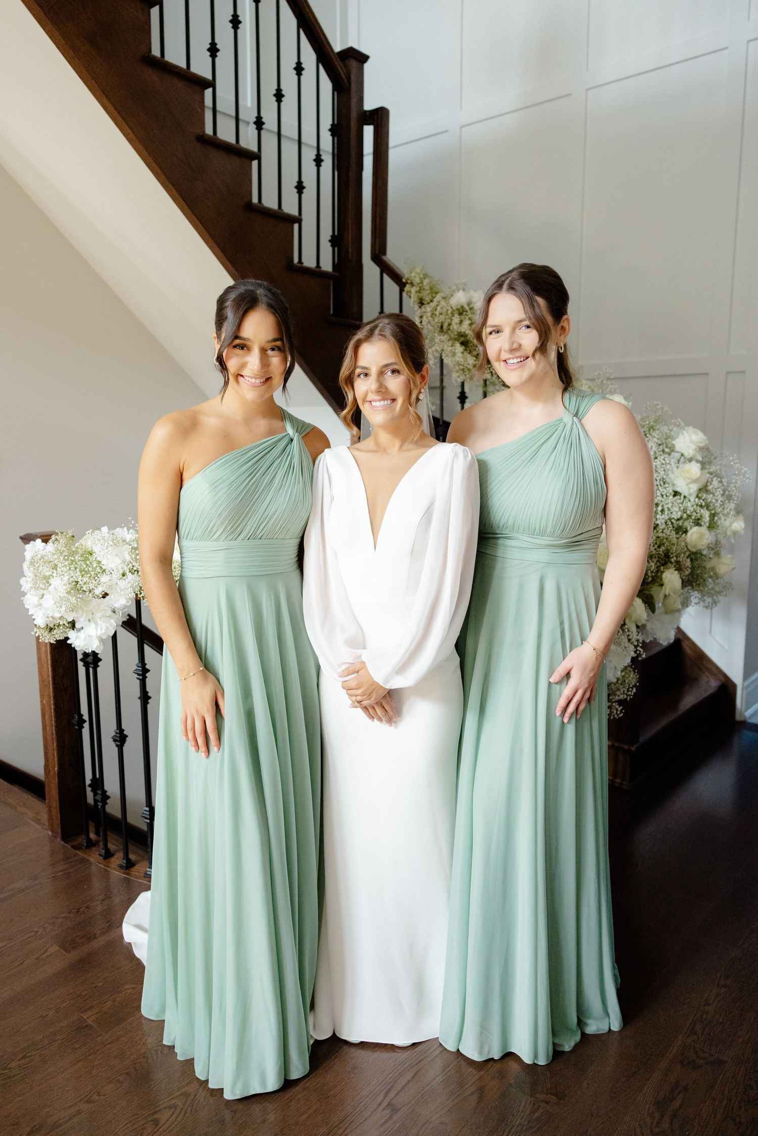Three women in coordinating mint green and white formal dresses pose by a wooden staircase.