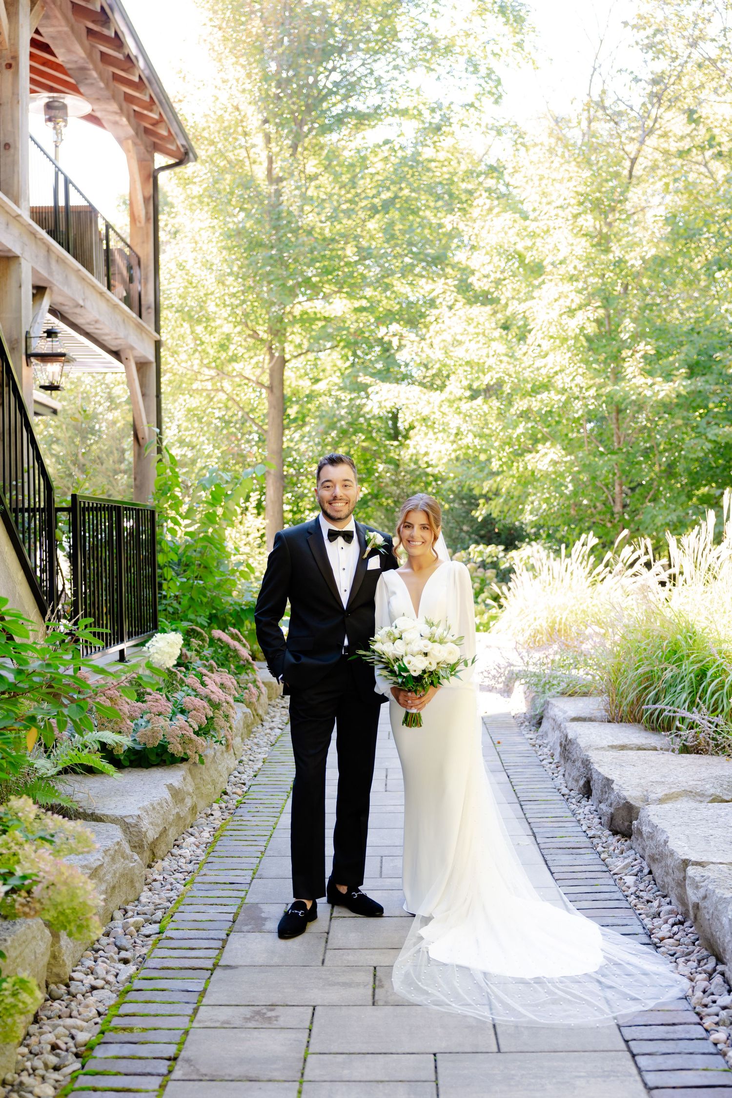 Wedding couple poses on landscaped stone pathway surrounded by lush summer foliage.