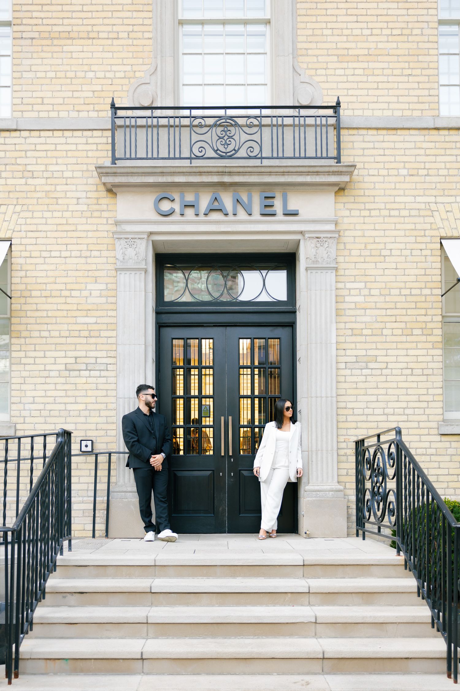 A couple stands on the steps of a Chanel boutique with cream brick facade.