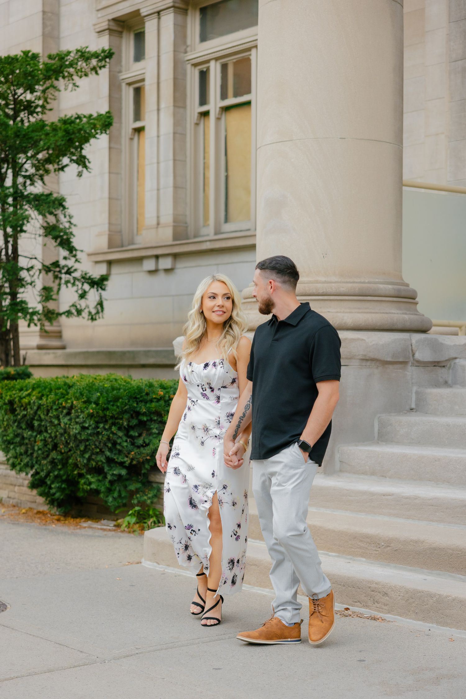 A couple stands together on a sidewalk next to stone columns and manicured shrubs.