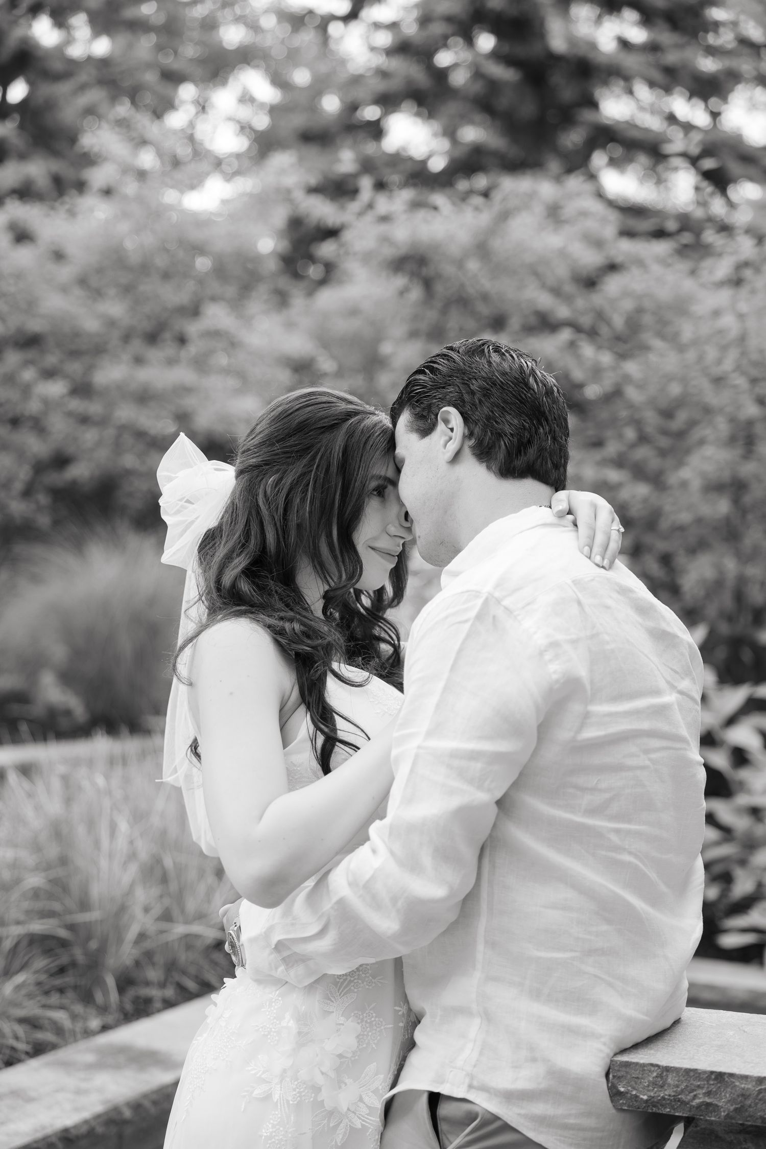 Black and white romantic wedding photo of a couple sharing a tender moment on a stone bench.