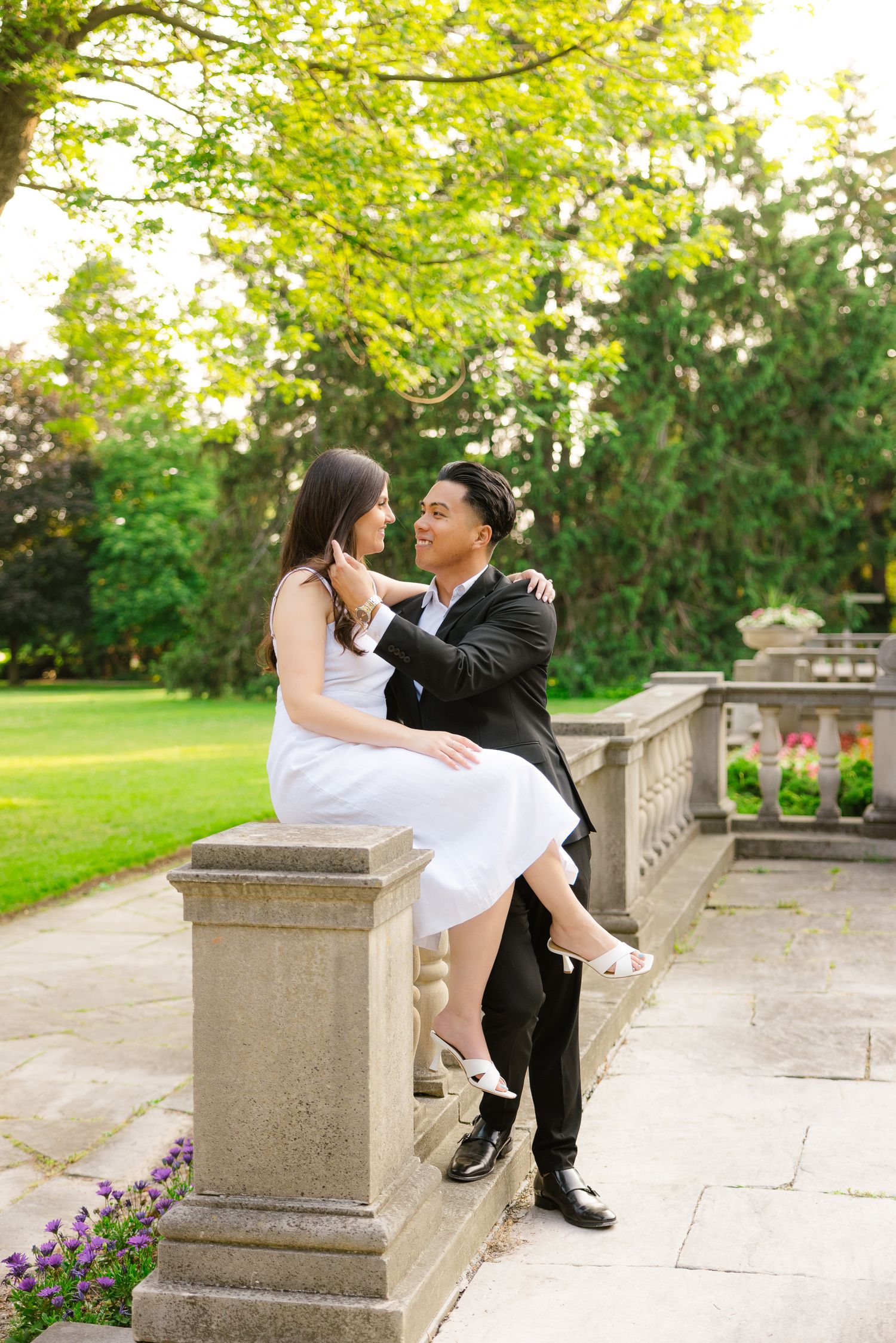 Couple shares intimate moment on stone railing with sunlit trees and garden in background.