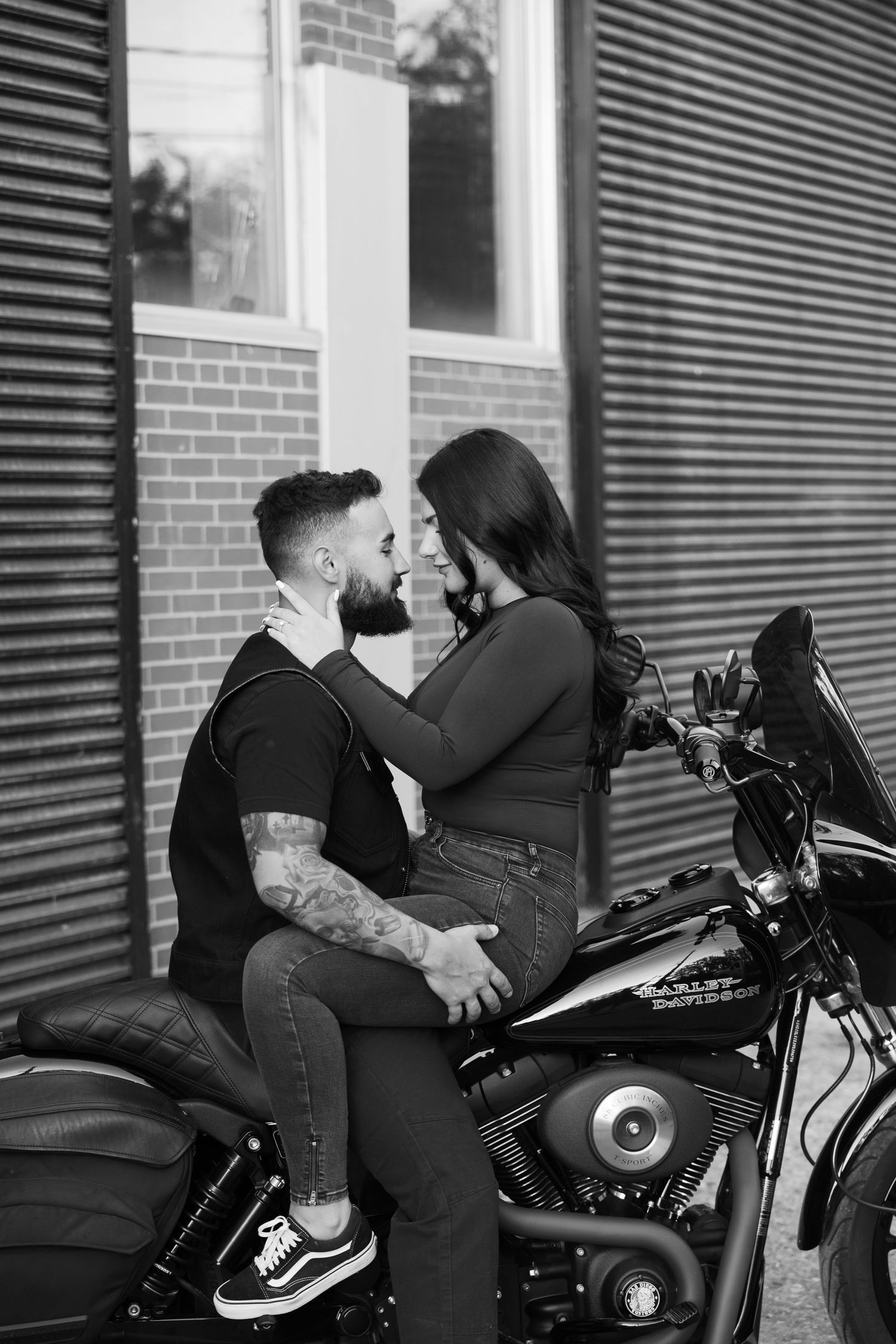 A couple embraces on a motorcycle in front of an industrial building.