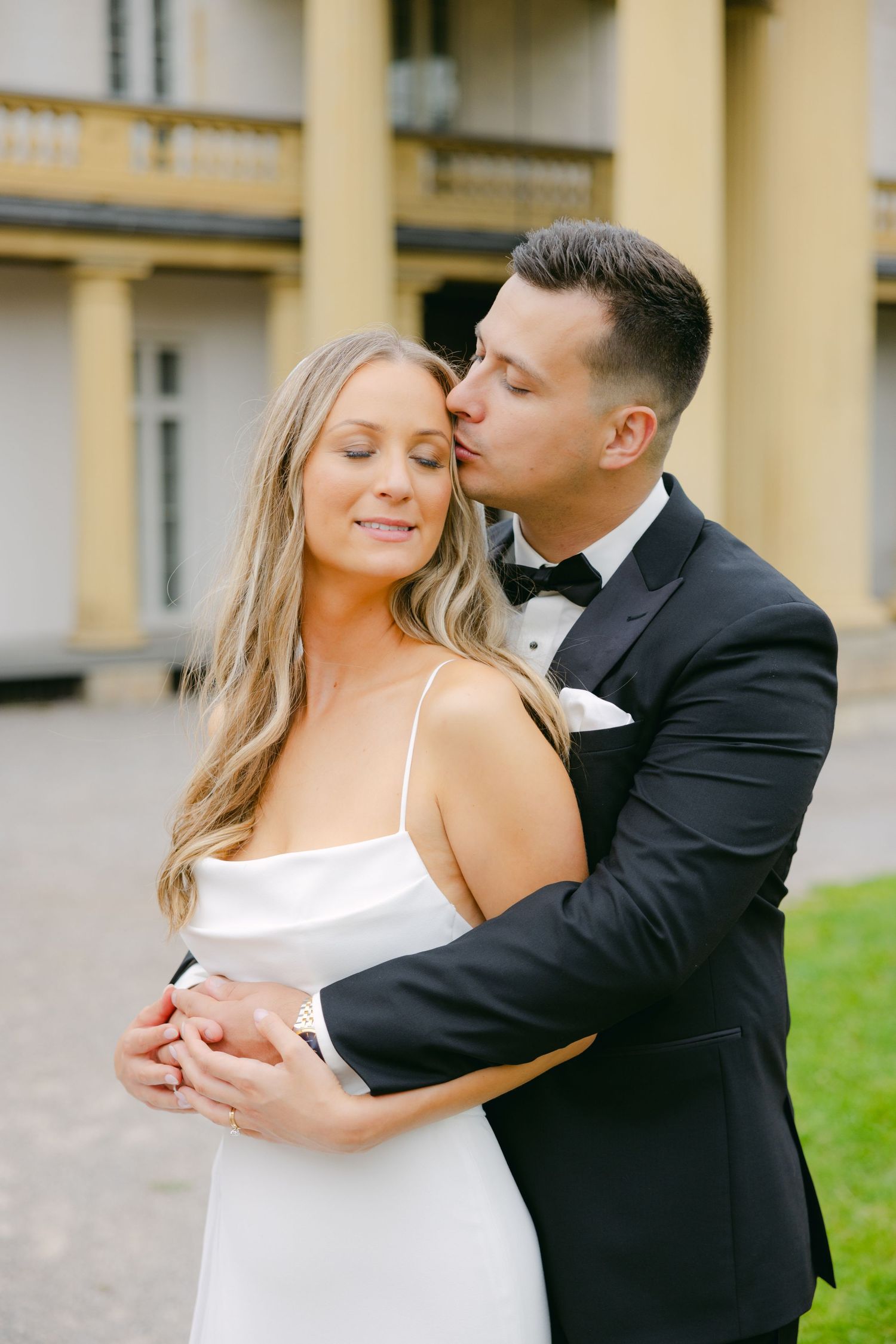 Tender moment between newlyweds against yellow mansion exterior.