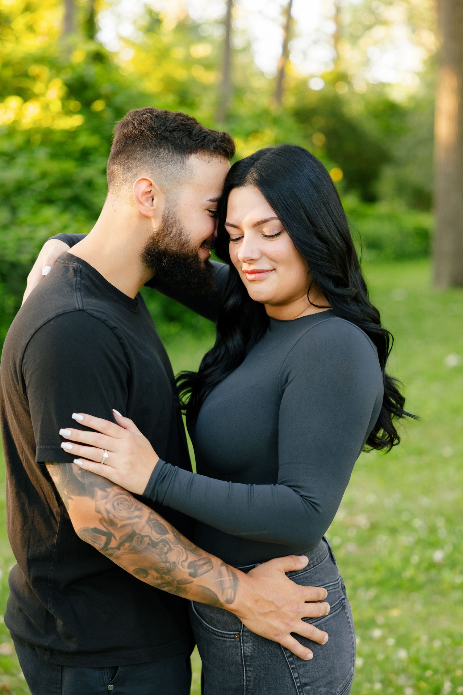 A couple in dark clothing shares an intimate moment in a sun-dappled forest clearing.