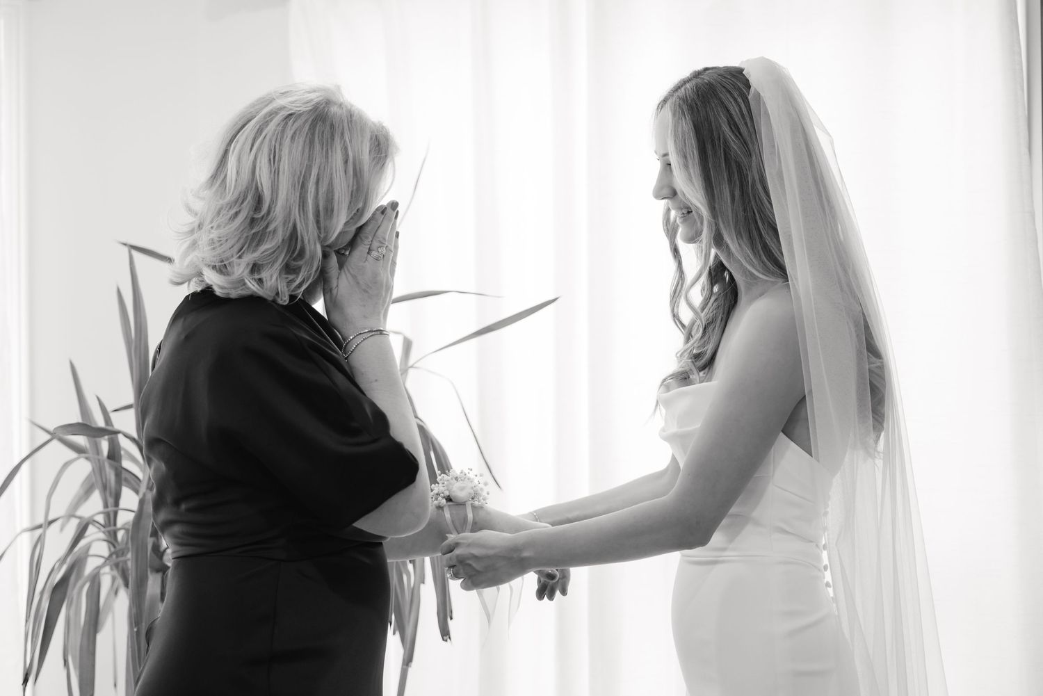 Tender moment between bride and older woman in black dress by bright window.