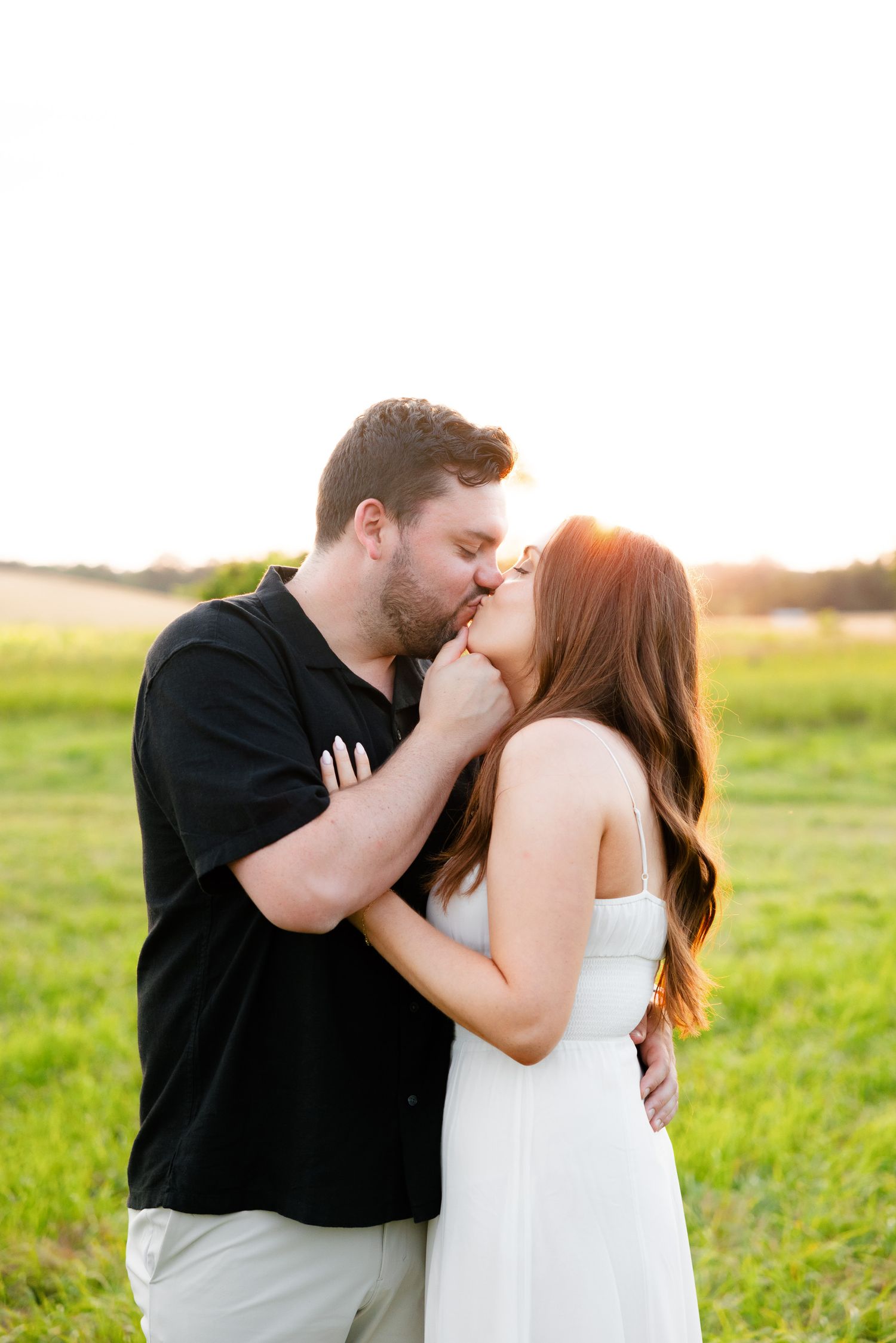 Couple shares romantic kiss in sunlit field wearing black and white outfits.