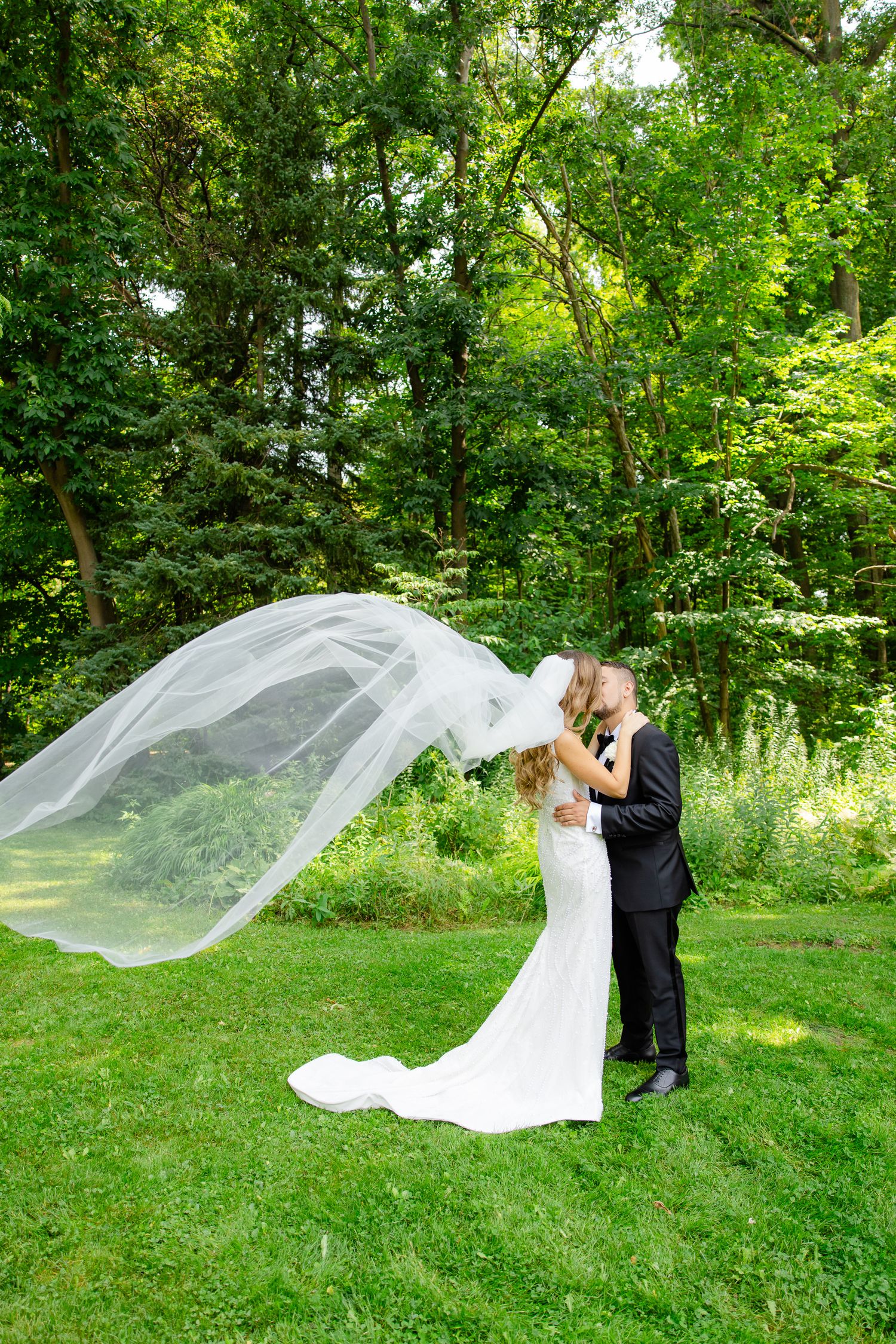 A stunning wedding portrait with a billowing veil catching the breeze in a green garden.