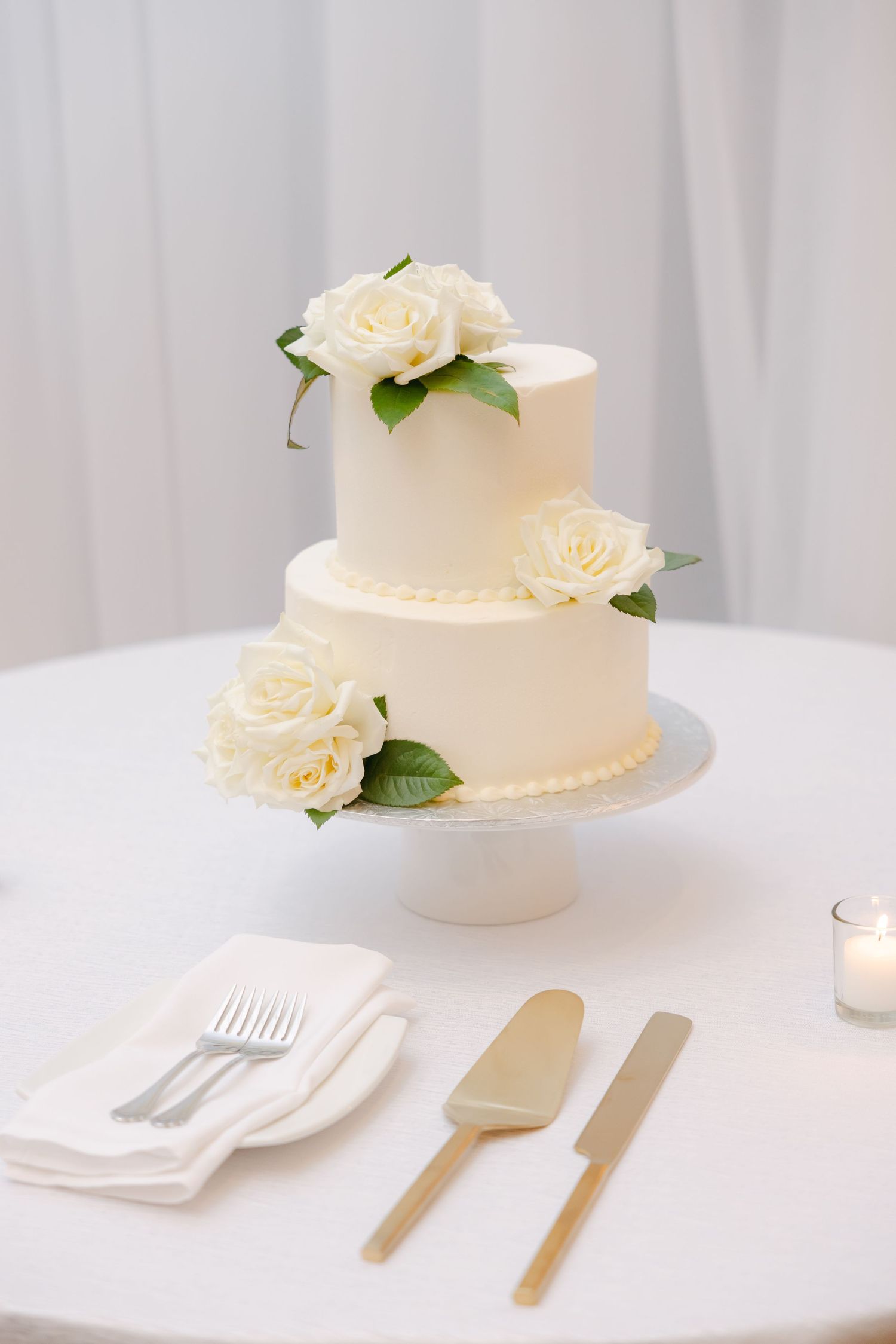 Two-tier white wedding cake decorated with fresh cream roses and gold serving utensils.