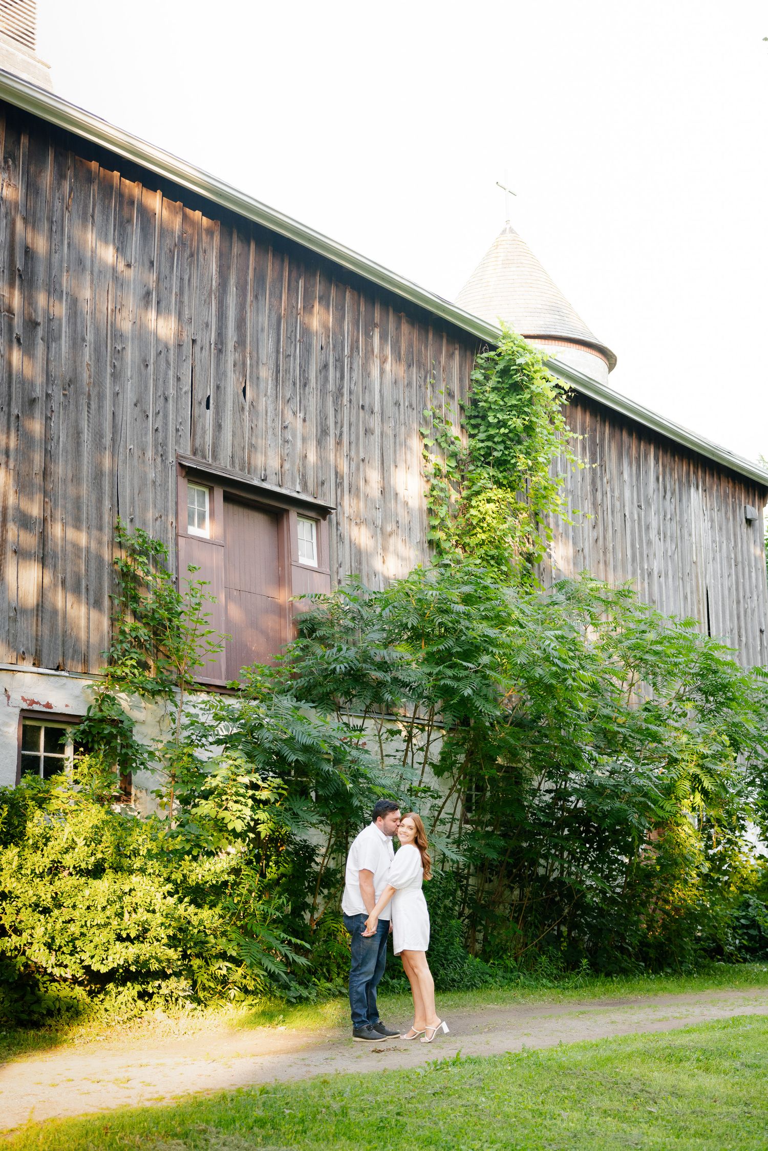 A couple embraces near a historic barn with weathered wood siding and climbing greenery.