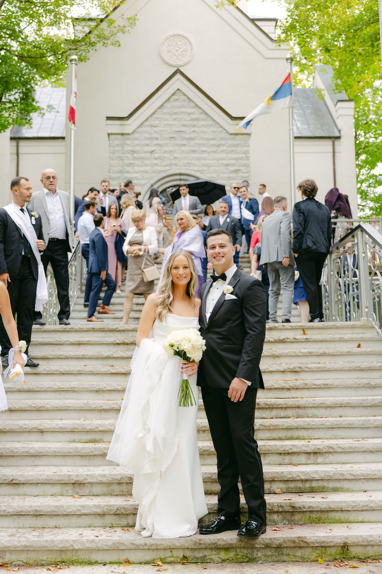 A wedding couple poses on church steps while guests gather in the background.