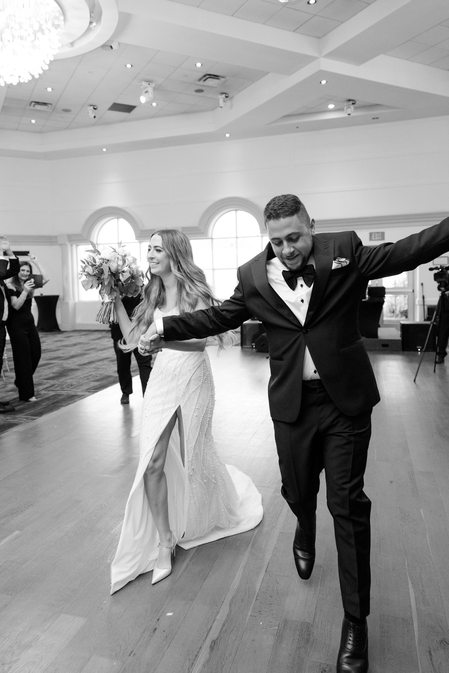 Joyful couple dances energetically across elegant ballroom with white arched windows and hardwood floors.
