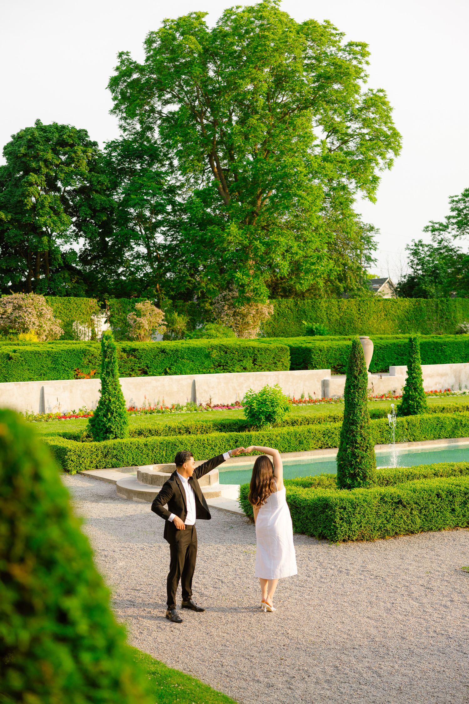 Two people share a playful moment on a garden path surrounded by manicured hedges and tall trees.