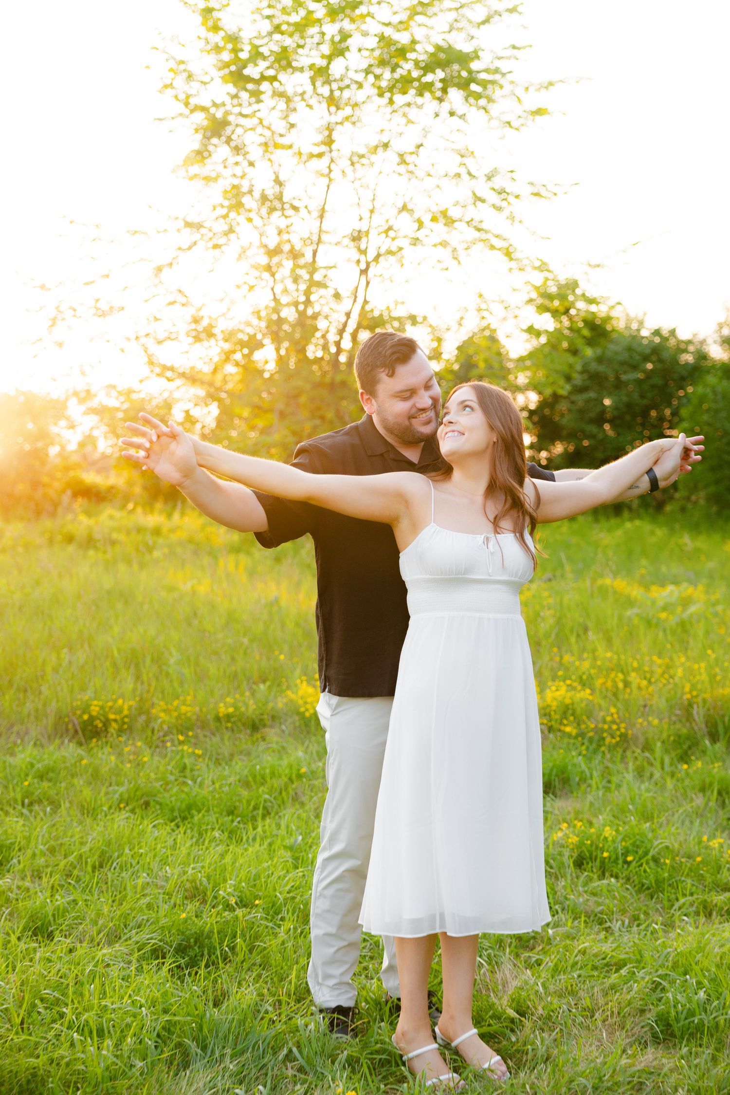 Joyful couple poses with arms outstretched in sunlit summer meadow.