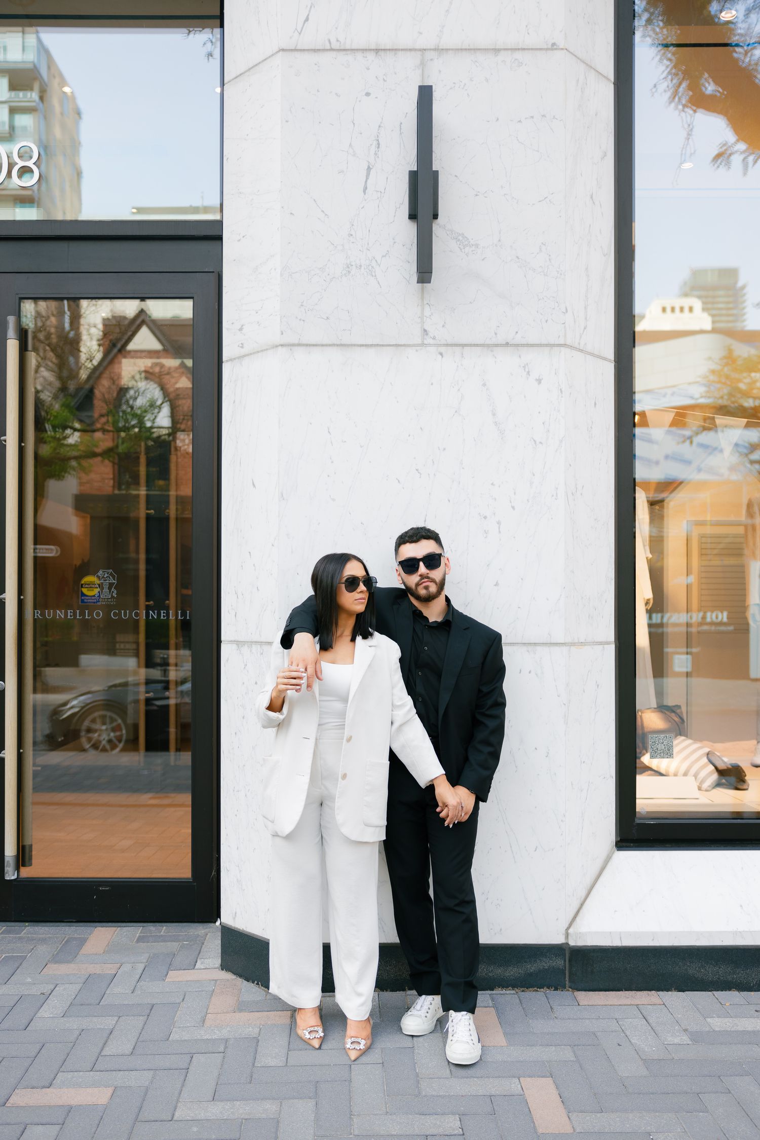 Couple in coordinated formal wear holding hands while standing against white storefront wall.
