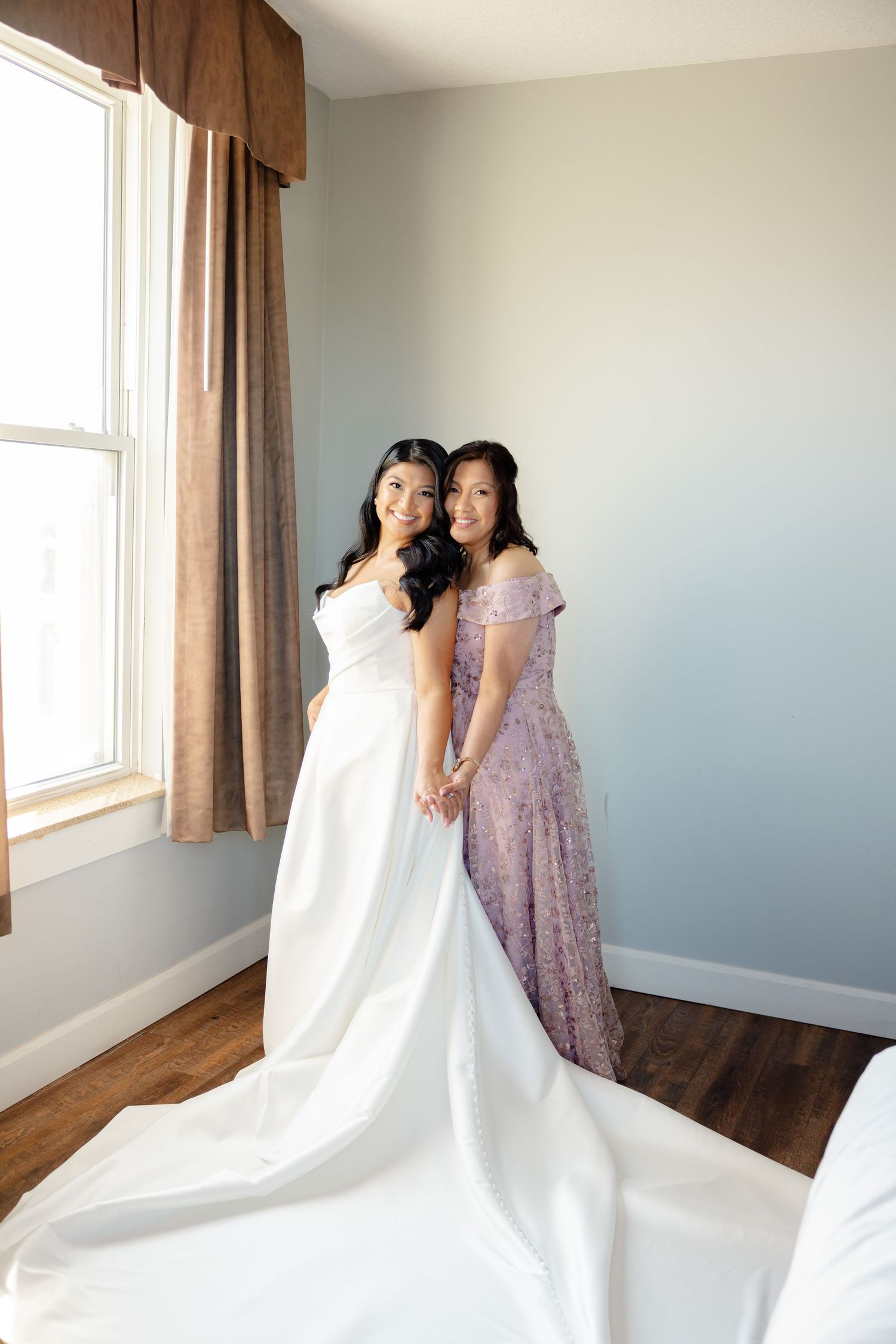 A tender pre-wedding moment captured near a window, featuring elegant white and lavender dresses.