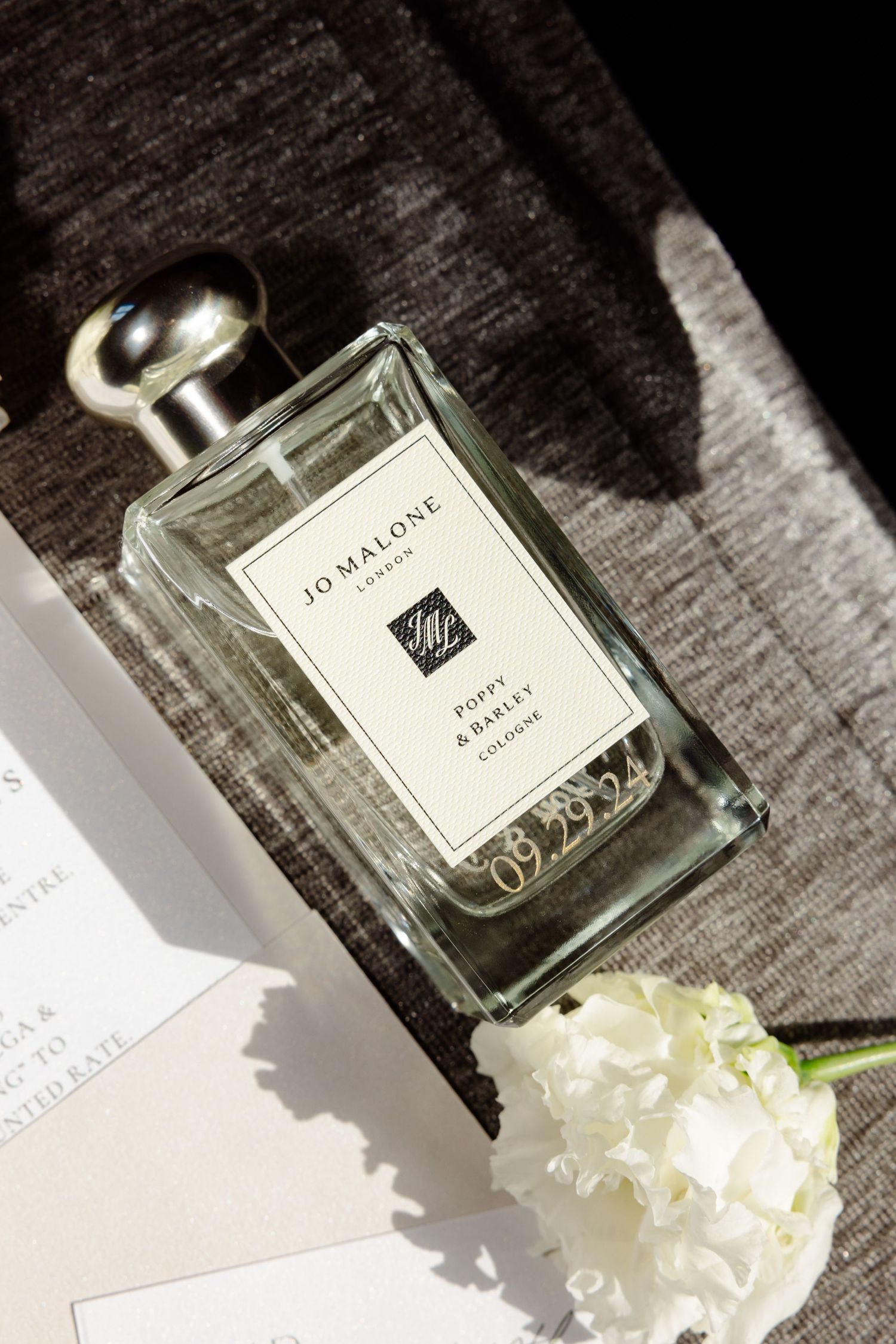 Jo Malone perfume bottle photographed with white flower on textured gray surface.