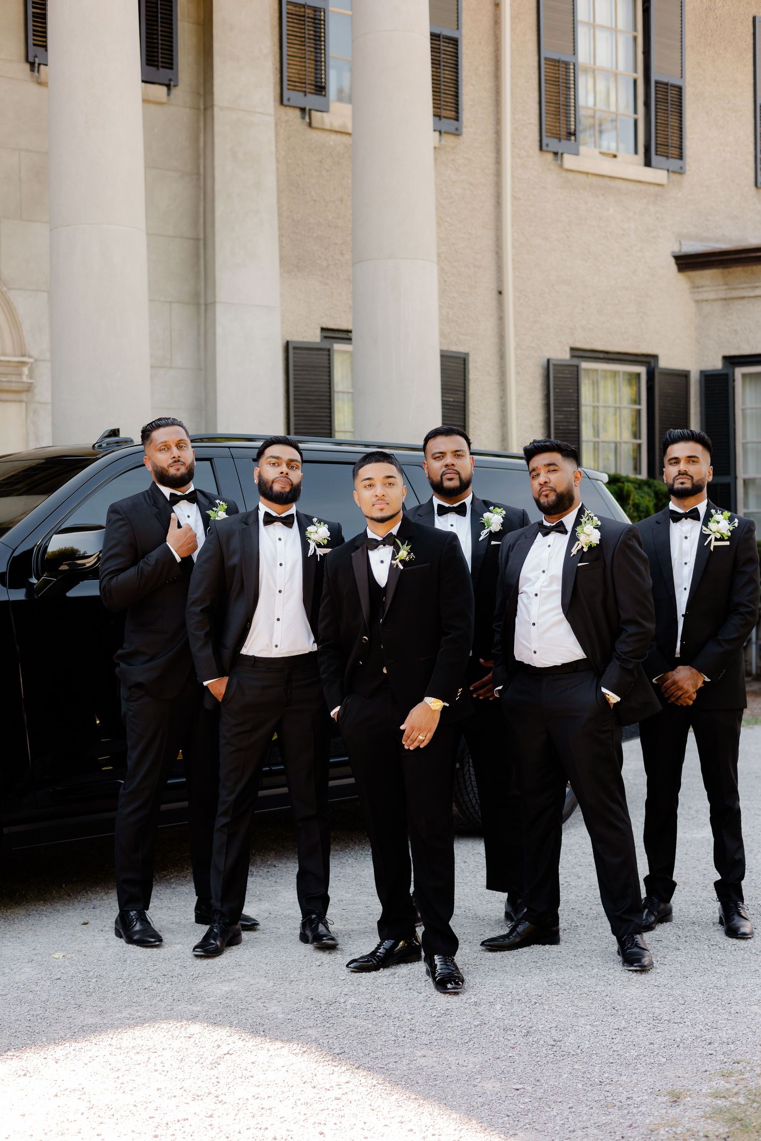 Groomsmen in black tuxedos pose in front of a luxury SUV and elegant building.