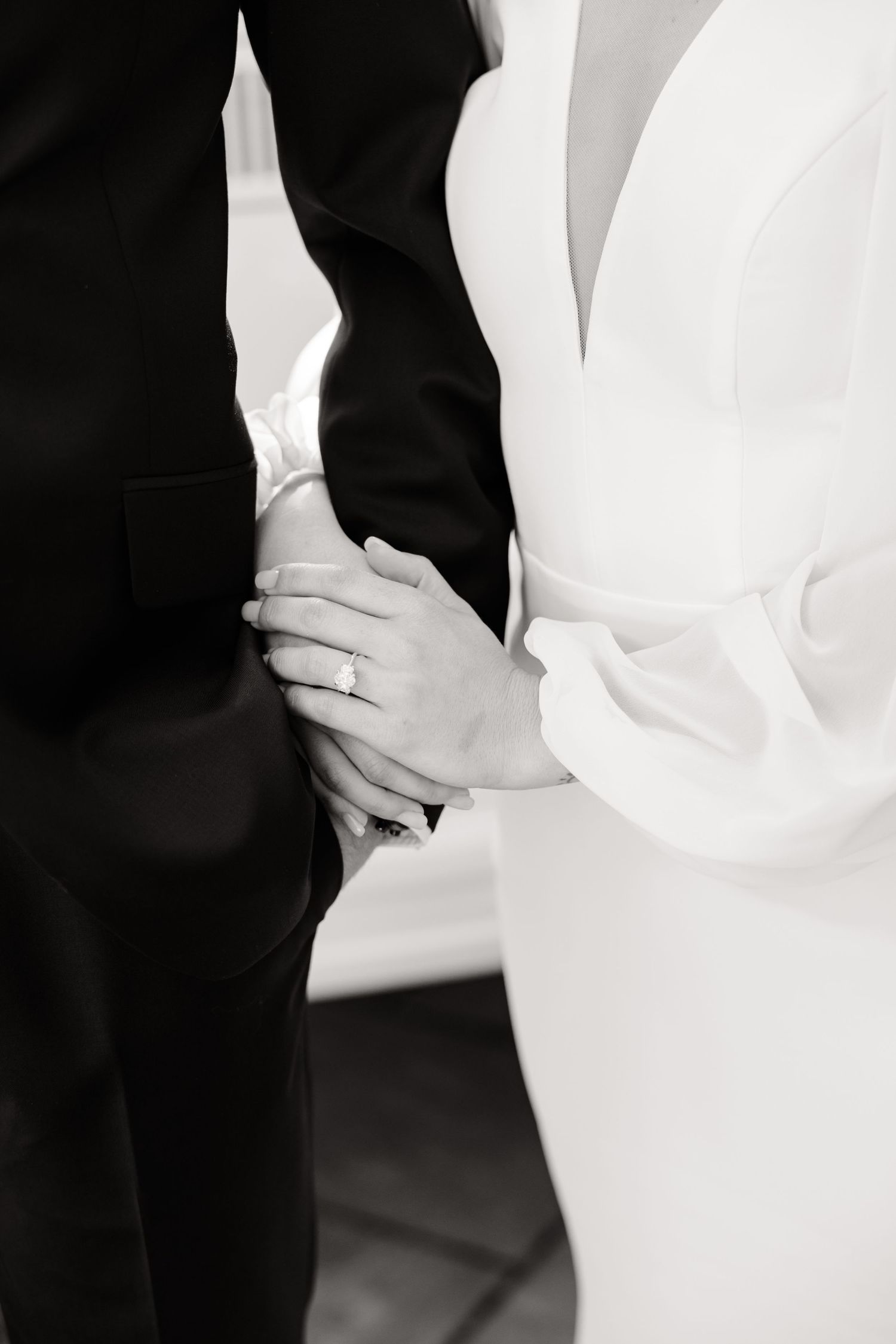 Close-up of wedding attire showing black tuxedo and white dress with hands clasped together.