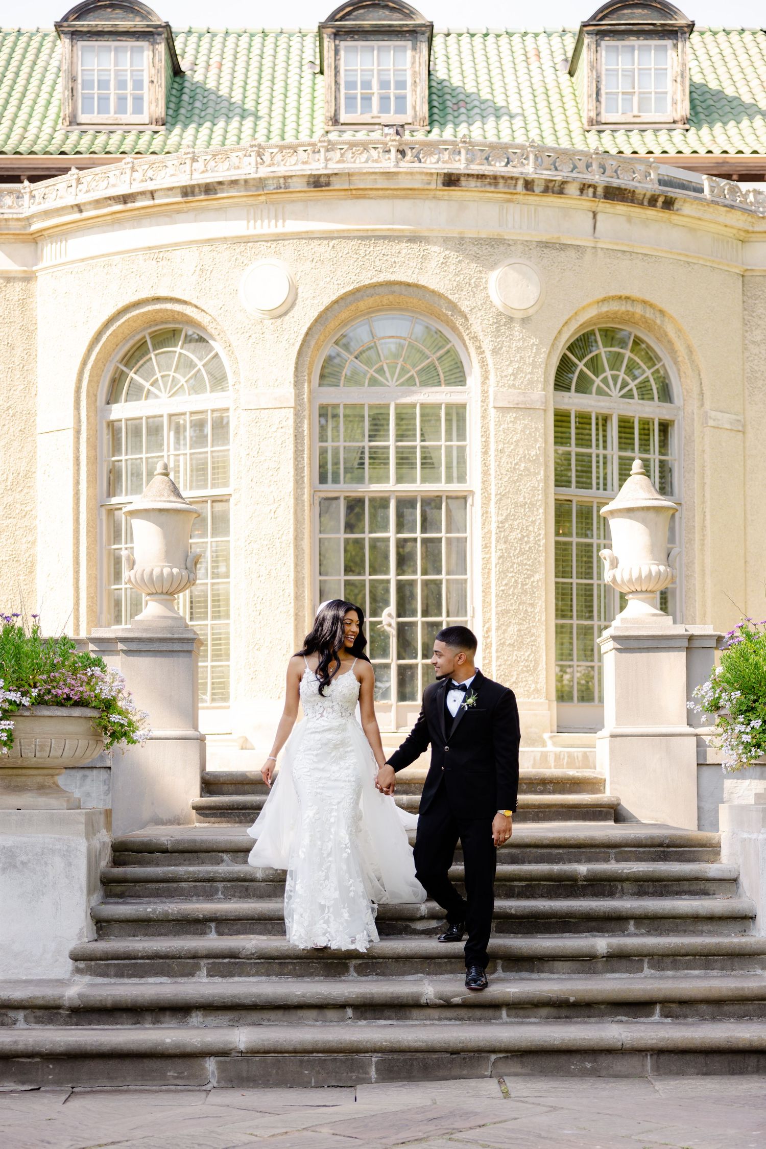 A couple descends elegant stone steps at a historic venue with tall arched windows and ornate architecture.