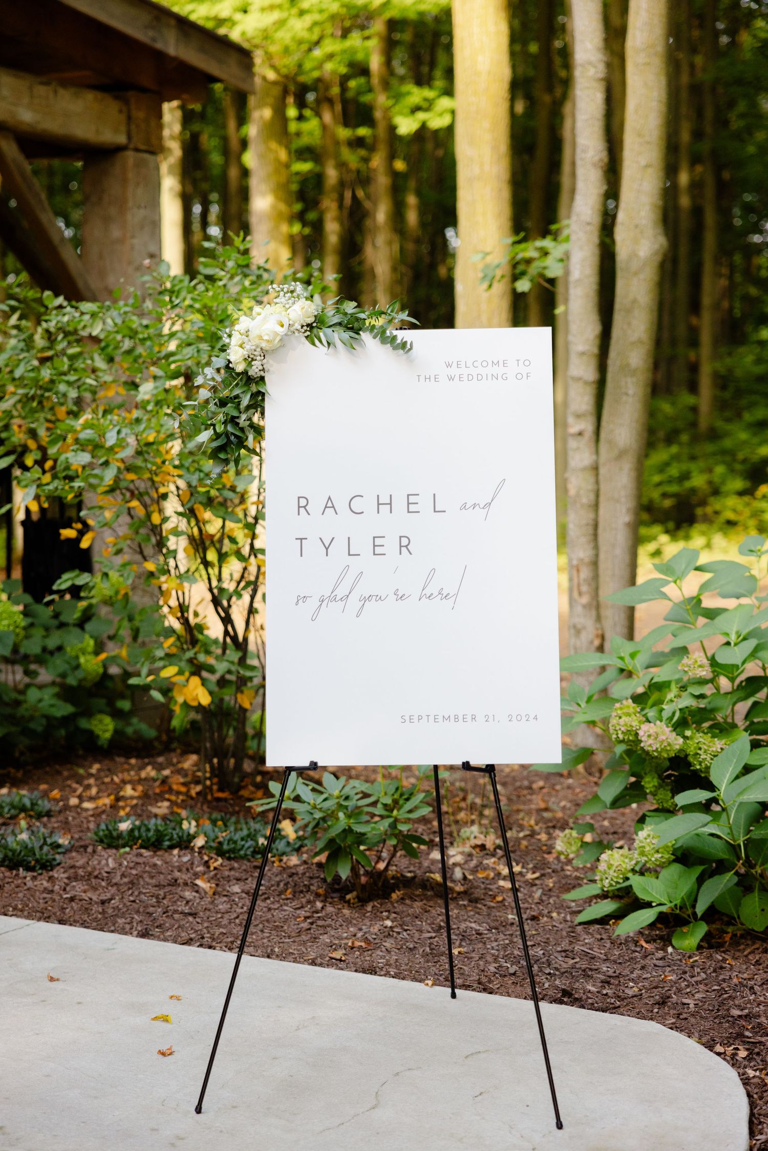 Wedding welcome sign with floral decor displayed on an easel in a wooded outdoor setting.