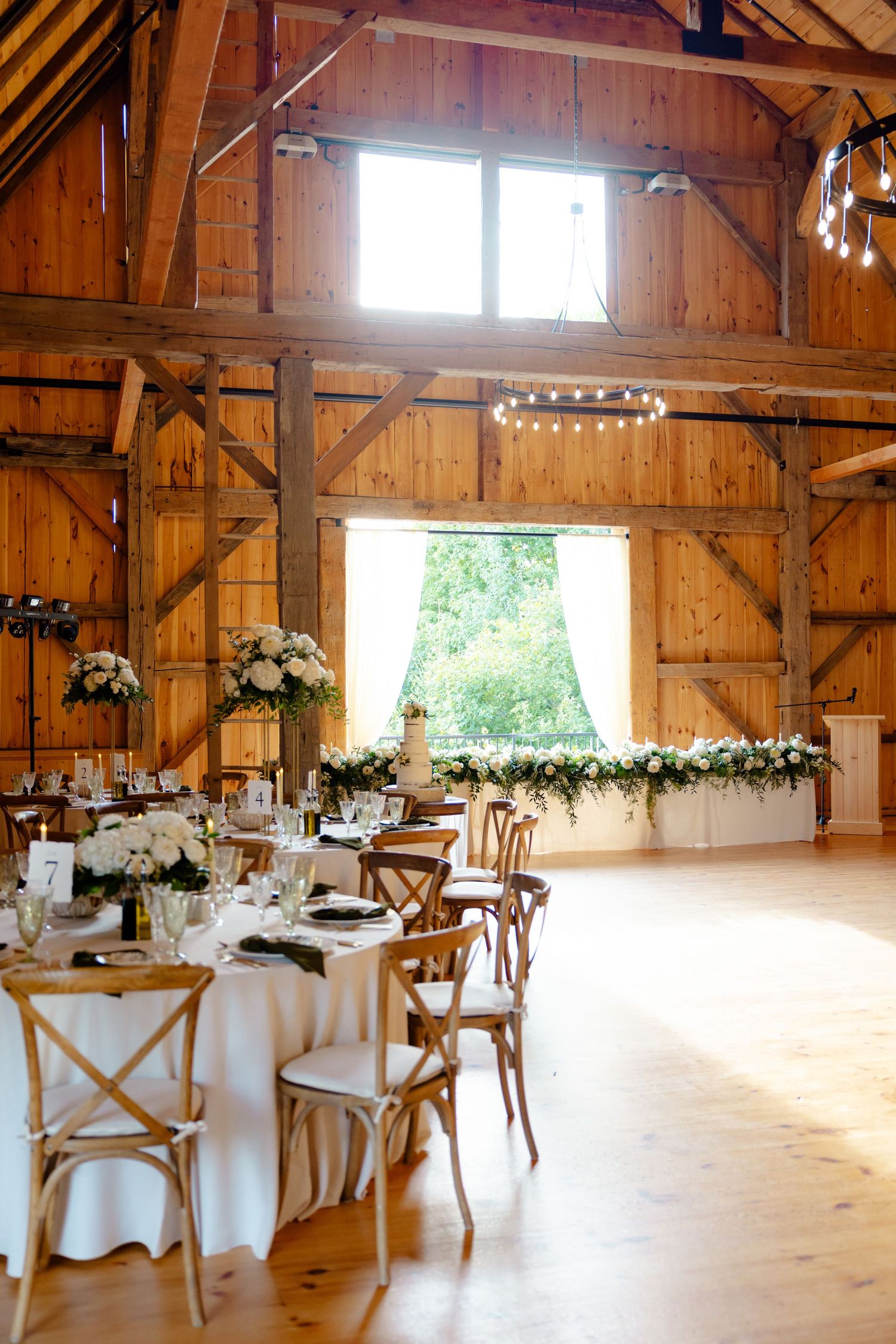 A sunlit barn reception hall with white tablecloths, cross-back chairs, and elegant window treatments.