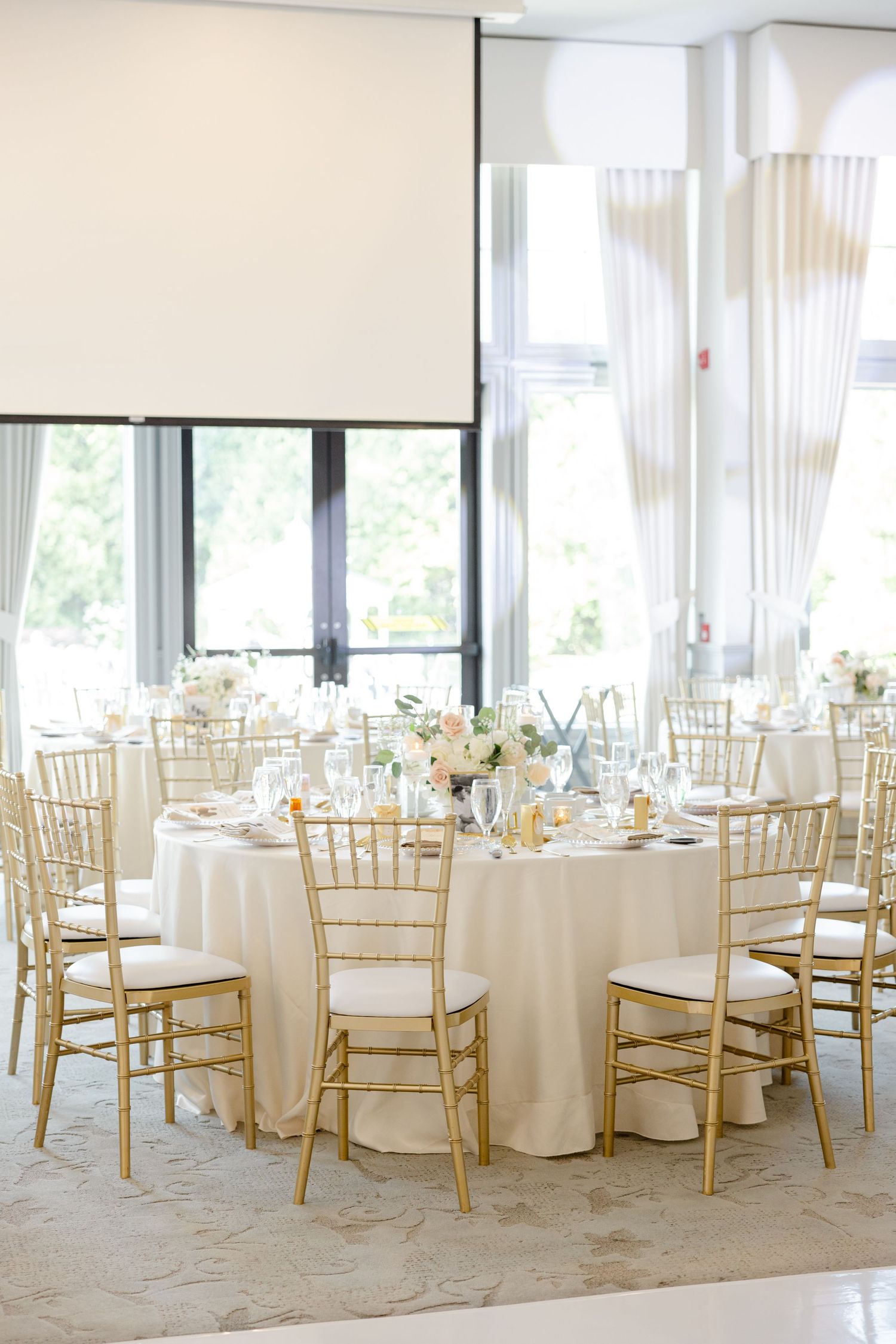 Bright wedding reception space with round tables, gold chairs, and cream-colored linens.