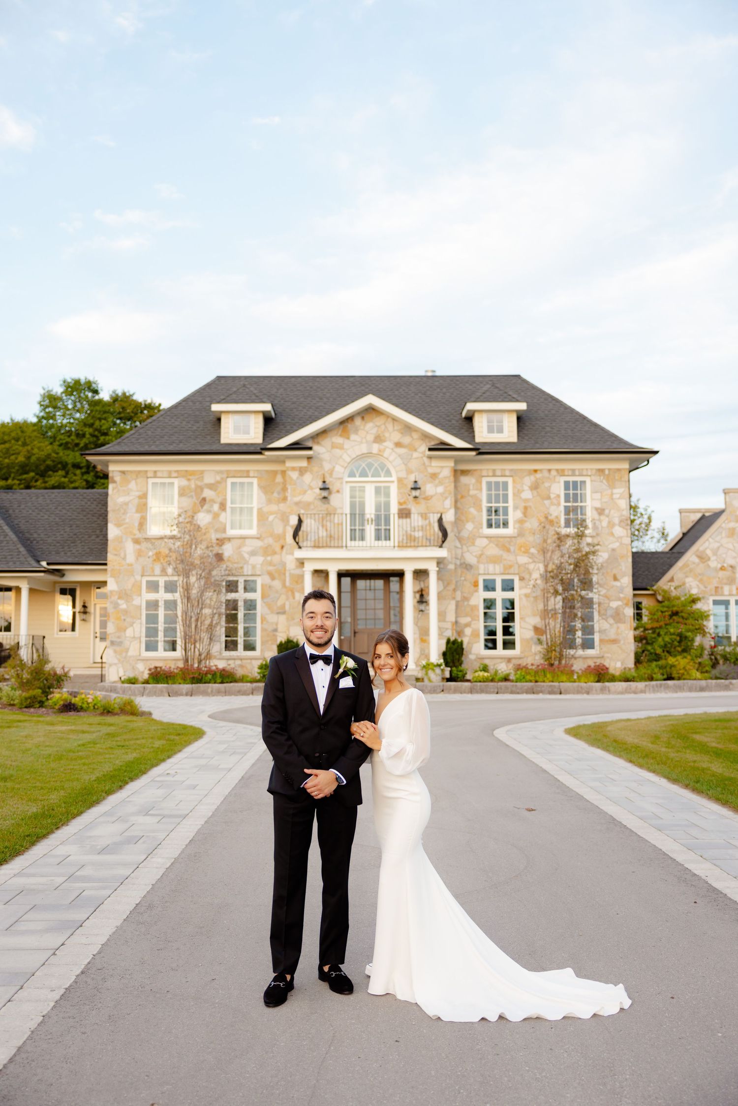 Newlyweds stand together on a driveway in front of an elegant stone mansion.
