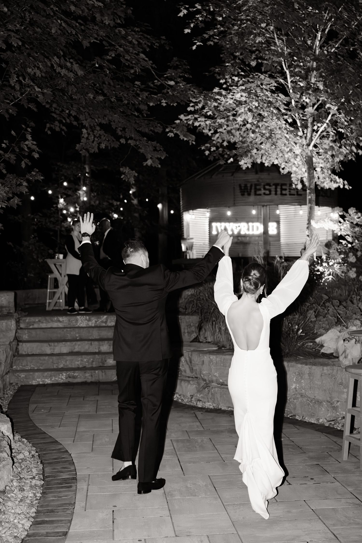 Black and white photo of wedding couple celebrating with arms raised on stone patio at night.