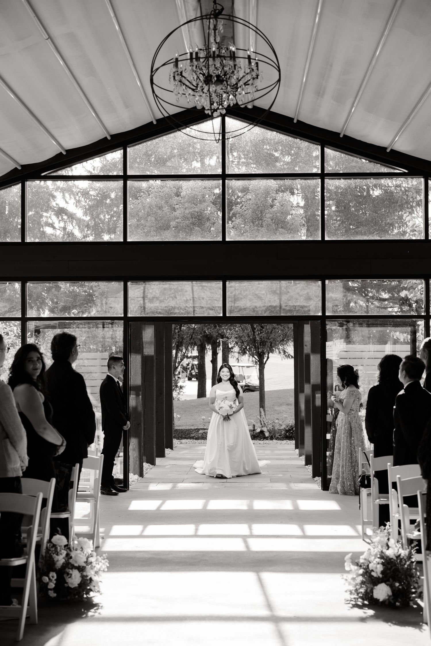 Black and white photo of a wedding ceremony in a modern glass venue with dramatic lighting.