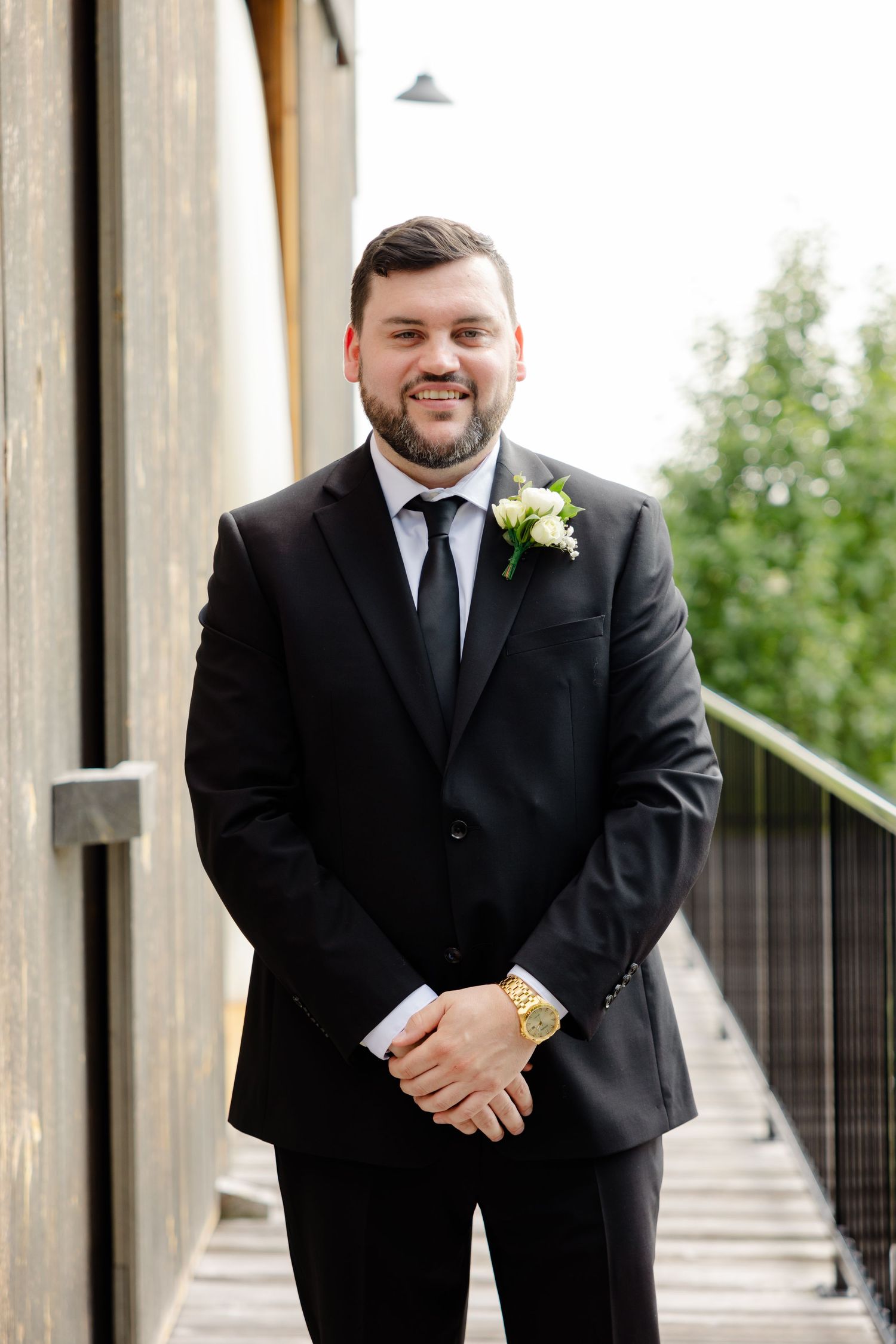 Person wearing black suit with boutonniere poses outdoors during formal event.