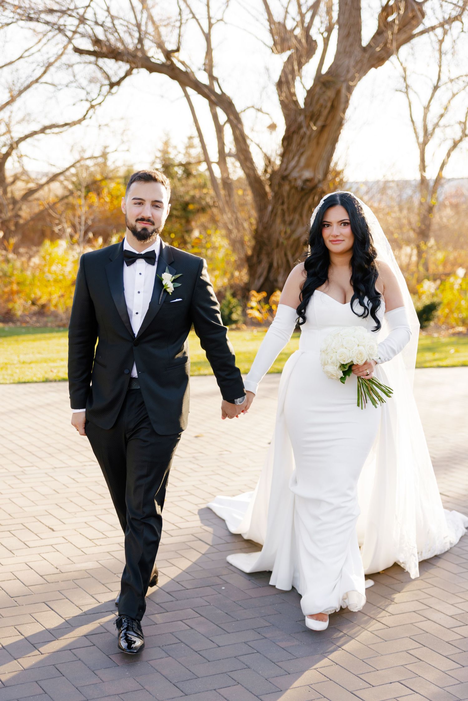 Wedding couple holding hands while walking on brick path with autumn scenery behind them.