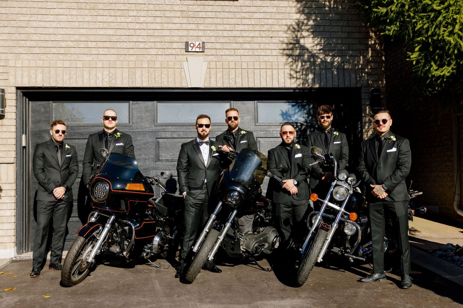 A group in black leather jackets poses with motorcycles in front of a garage door.